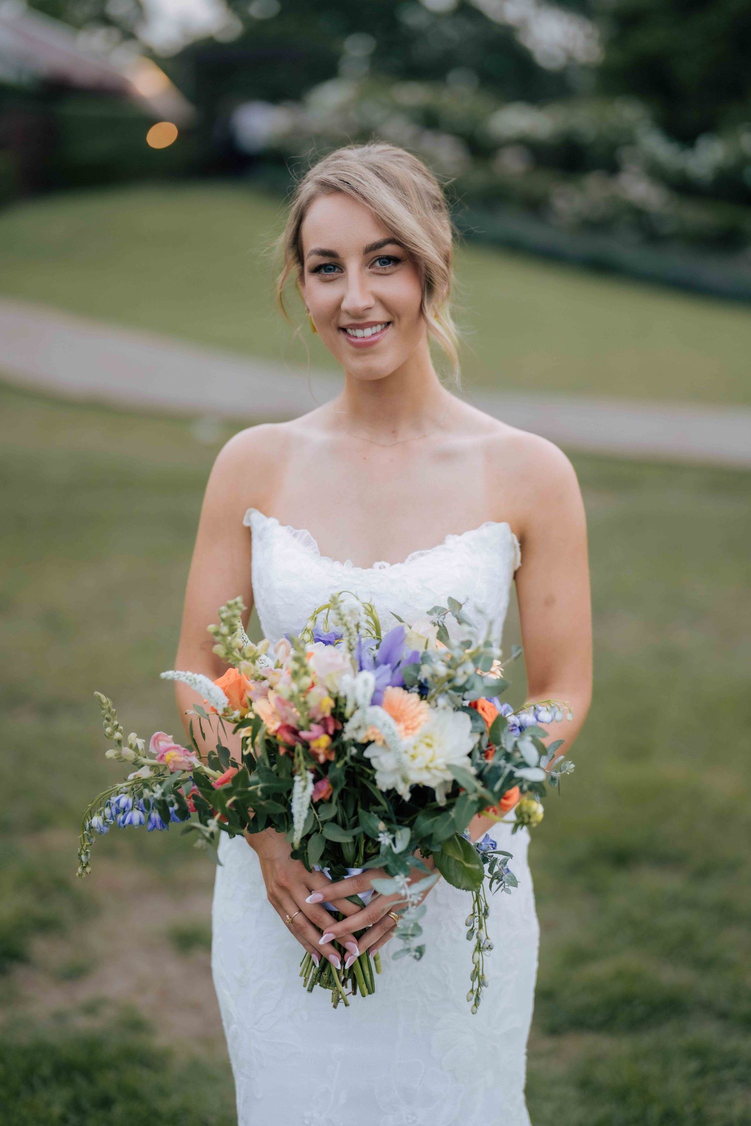 Elegant bride in strapless white lace gown smiles while holding a colorful spring bouquet in a garden setting.