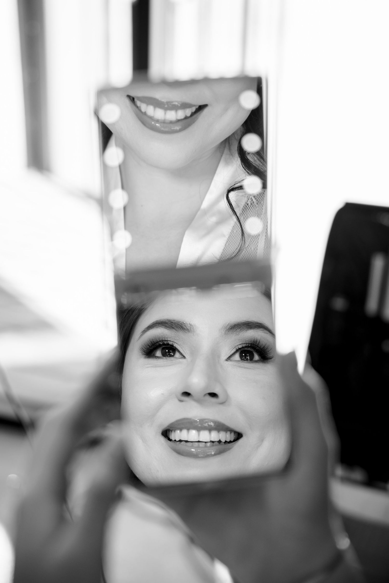 Bride smiles radiantly as her reflection appears in a handheld mirror during wedding preparations.