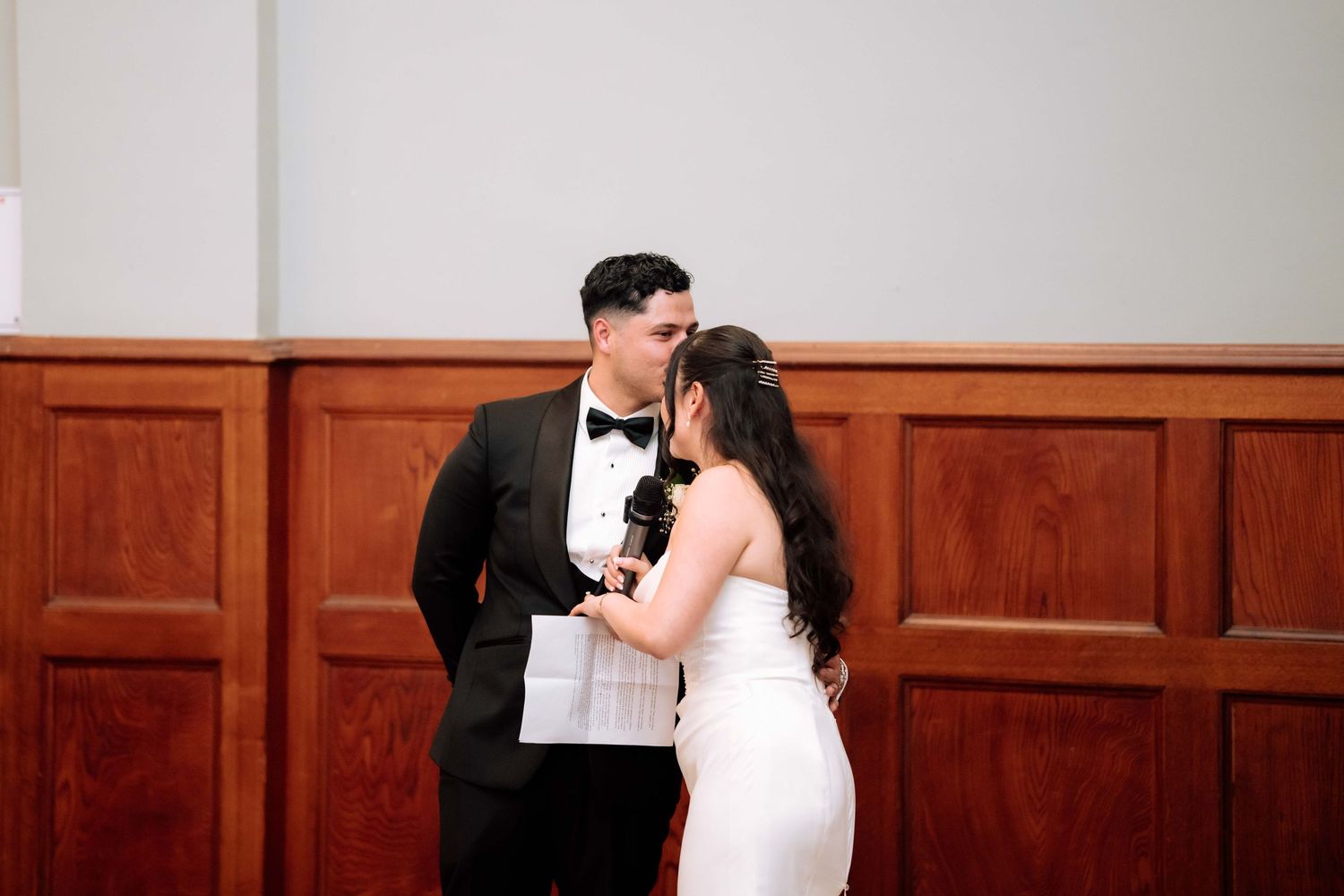 Bride and groom share a tender kiss while holding a microphone during their wedding reception speech.