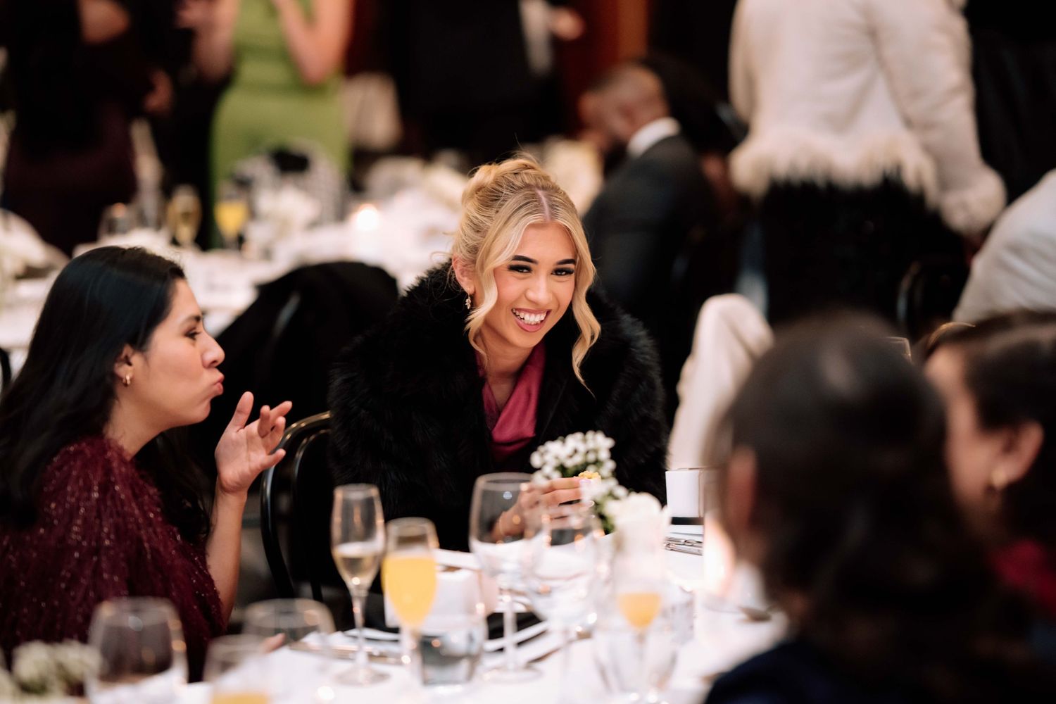 Smiling wedding guest in a fur stole laughing and chatting with others at a formal reception dinner.