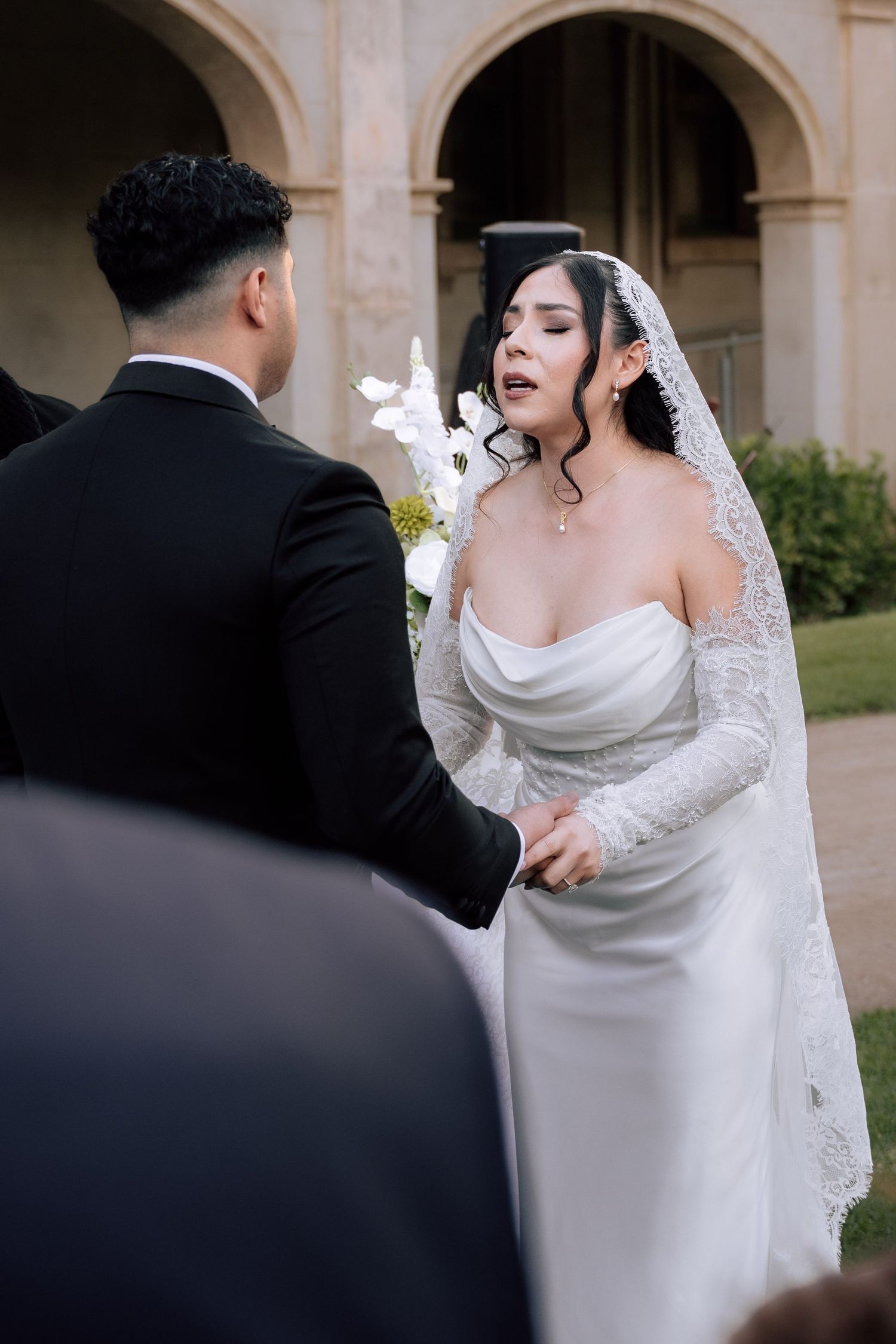 Bride in white strapless gown with lace veil exchanging vows with groom during outdoor wedding ceremony.