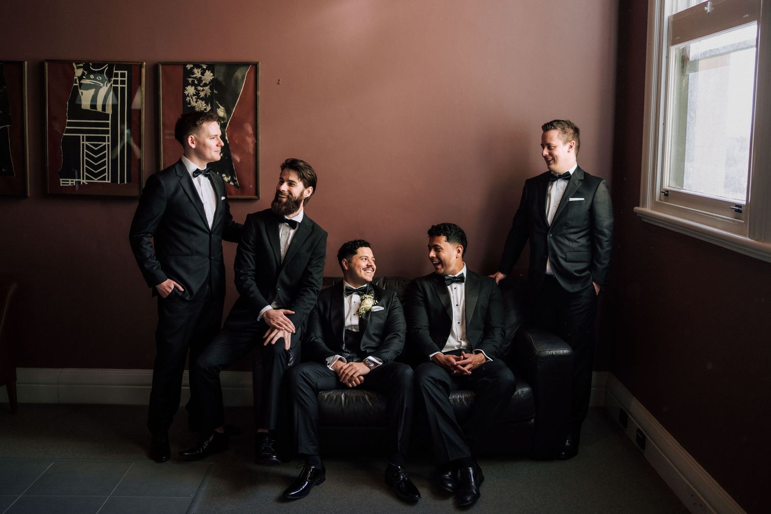 Five groomsmen in black tuxedos pose elegantly in a moody, warmly lit room before a wedding ceremony.