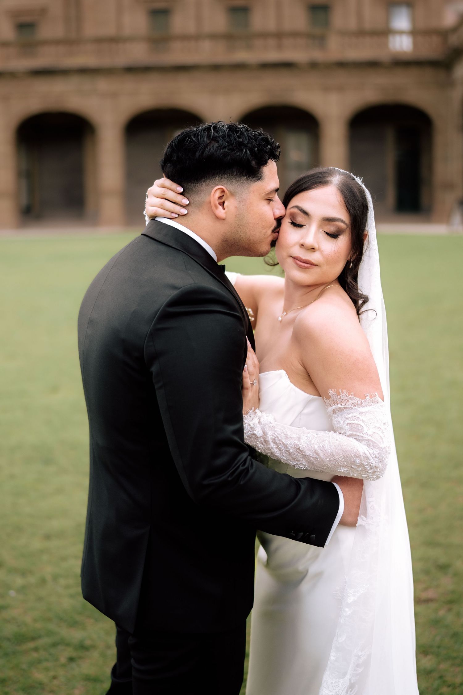 Groom in black tuxedo kissing bride in white lace off-shoulder gown outdoors at elegant venue.