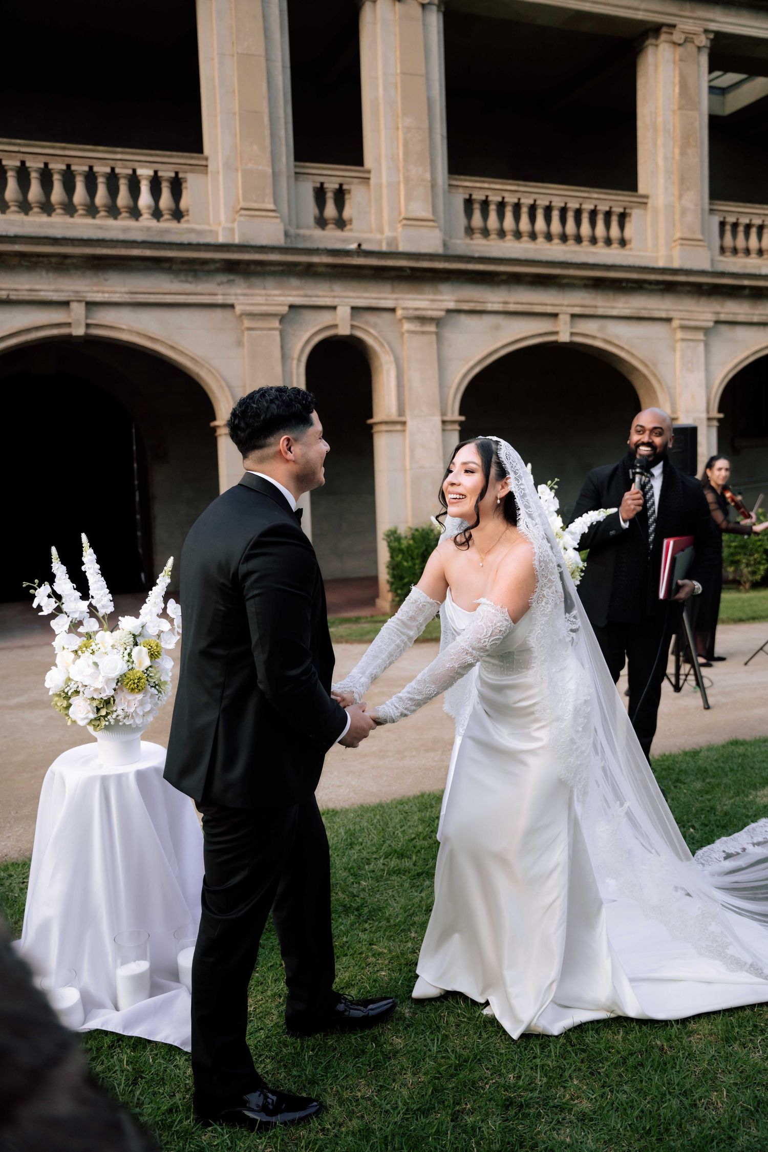 Bride in white gown and groom in black tuxedo hold hands during outdoor wedding ceremony at historic venue.