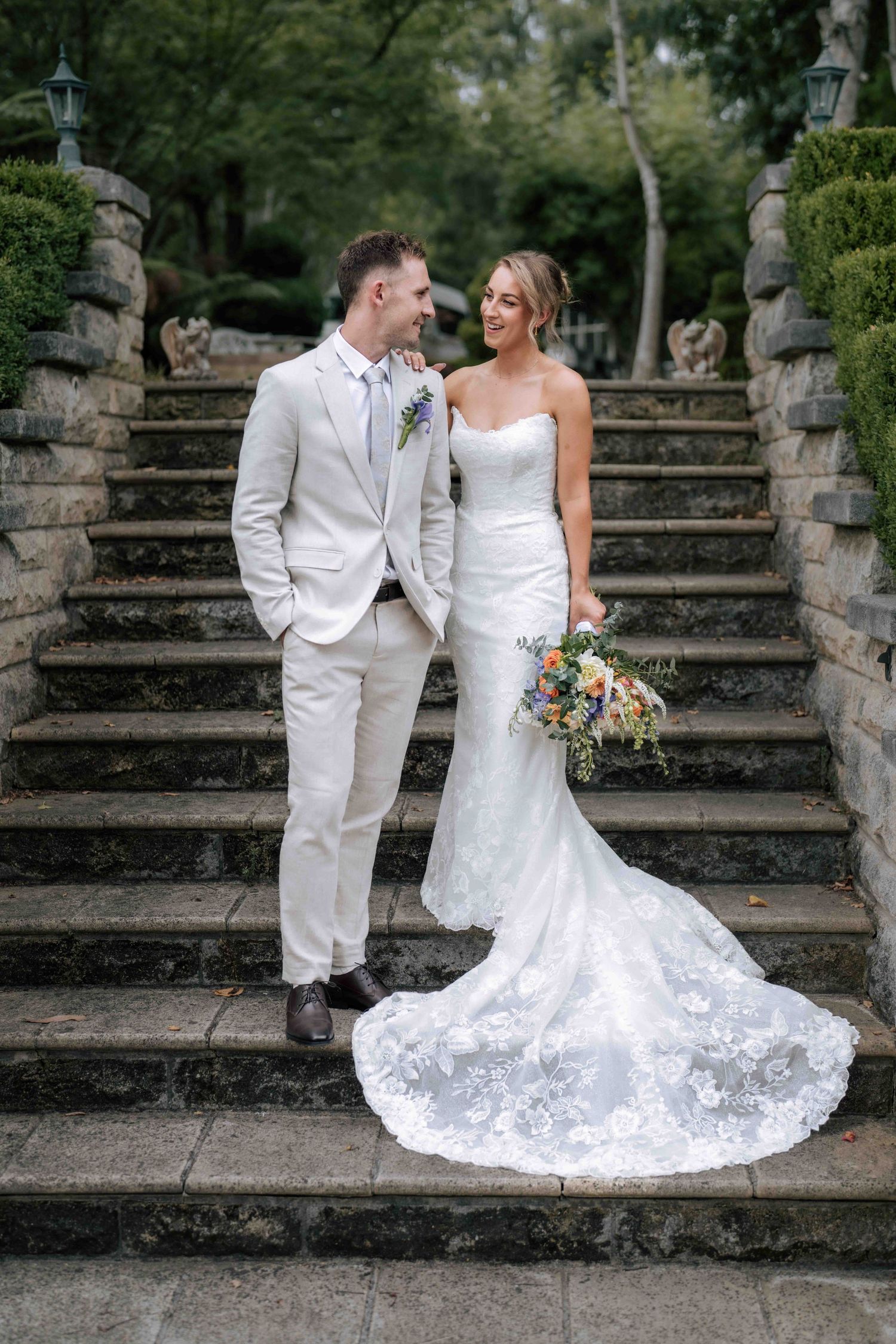 Bride and groom posing on stone steps, she in lace train gown holding bouquet, he in cream suit.