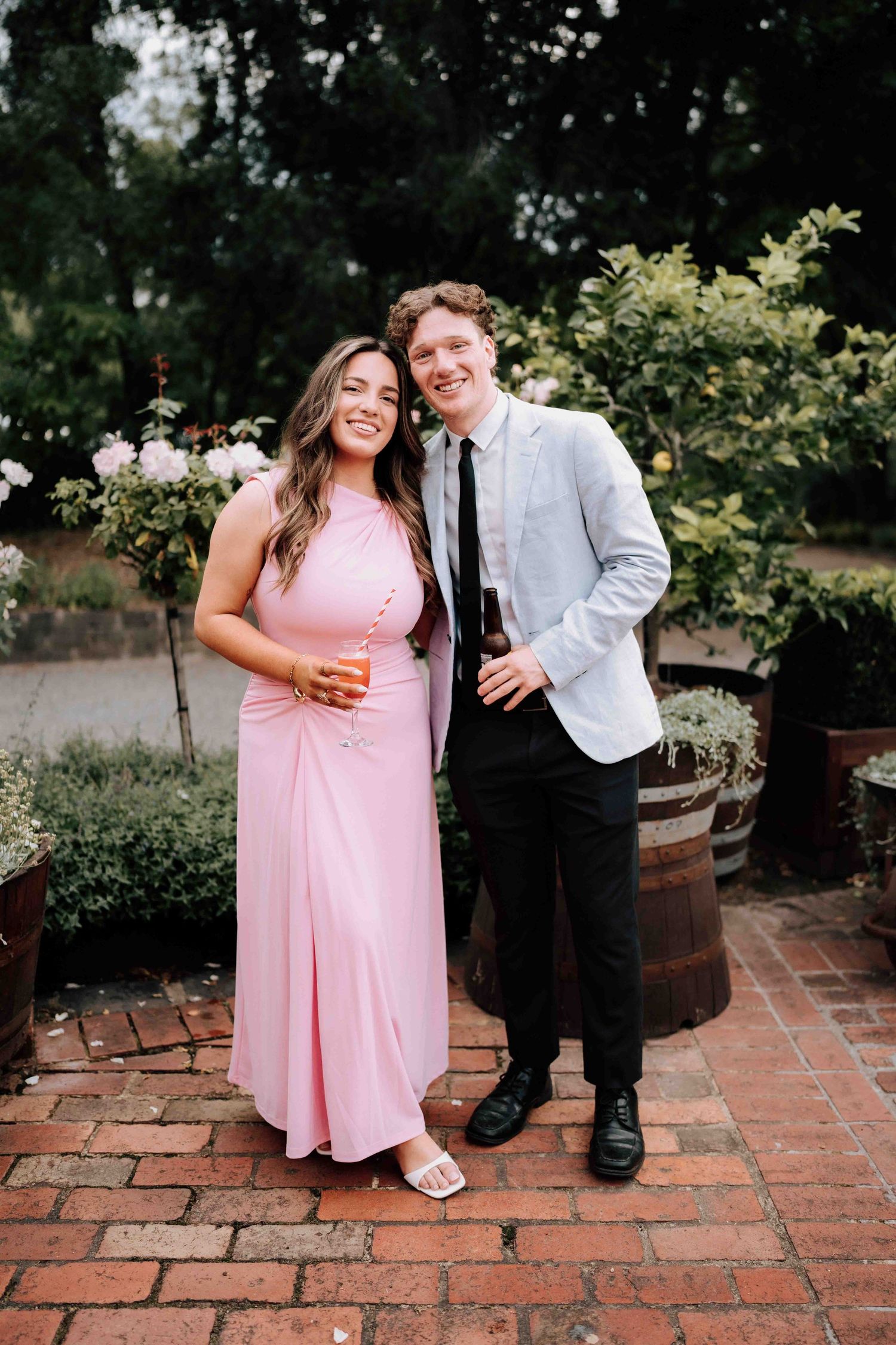 Smiling couple in pink dress and light blazer holding drinks in a garden at a wedding reception.