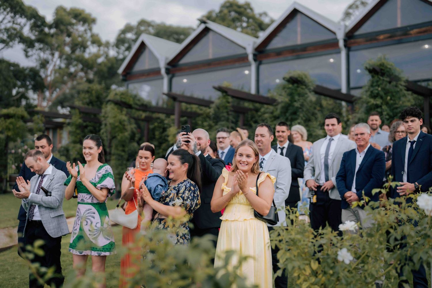 Wedding guests clapping and celebrating outdoors in front of modern venue building surrounded by lush gardens.