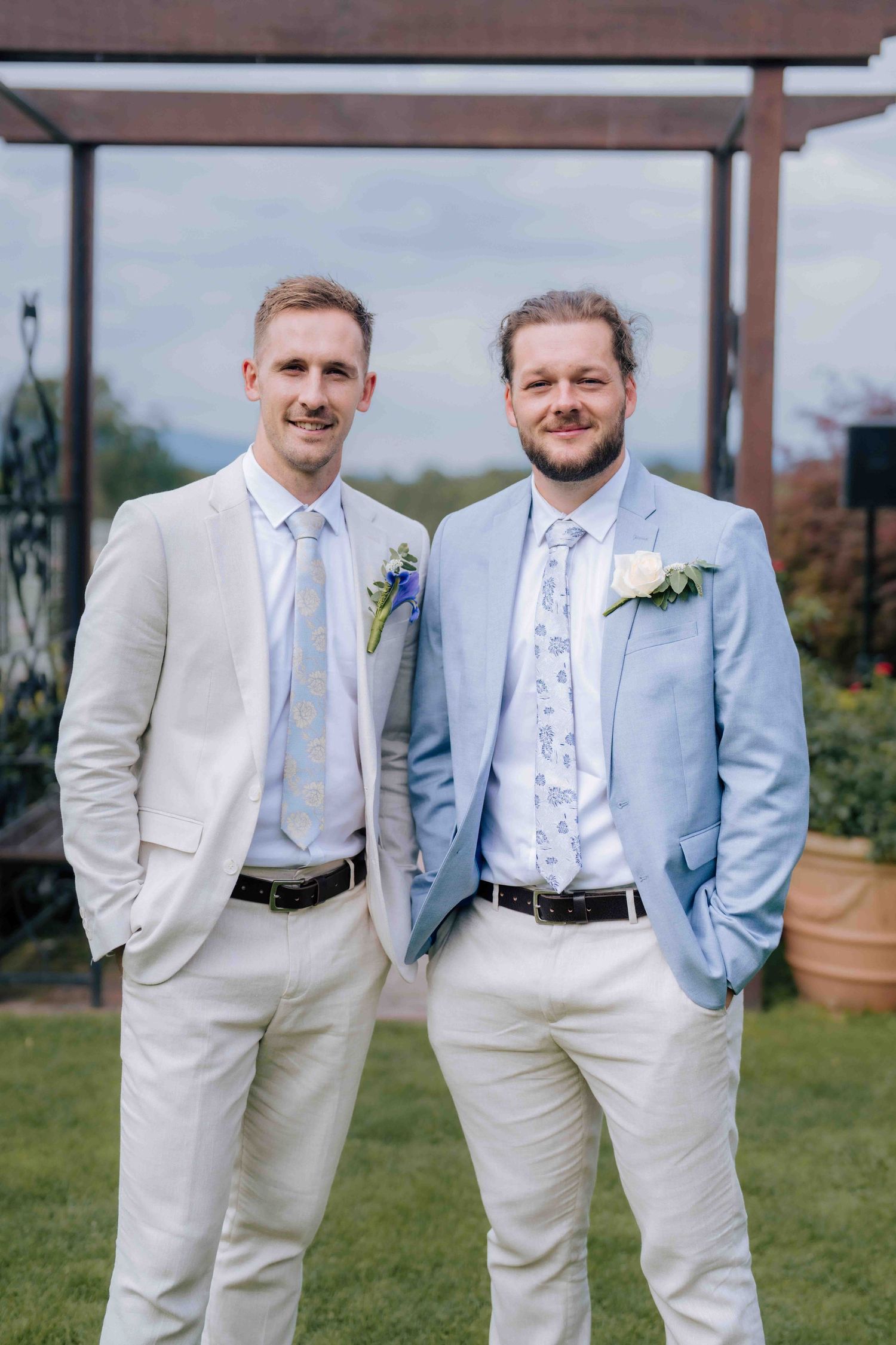 Two groomsmen in light suits with floral ties and white boutonnieres smiling at outdoor wedding.