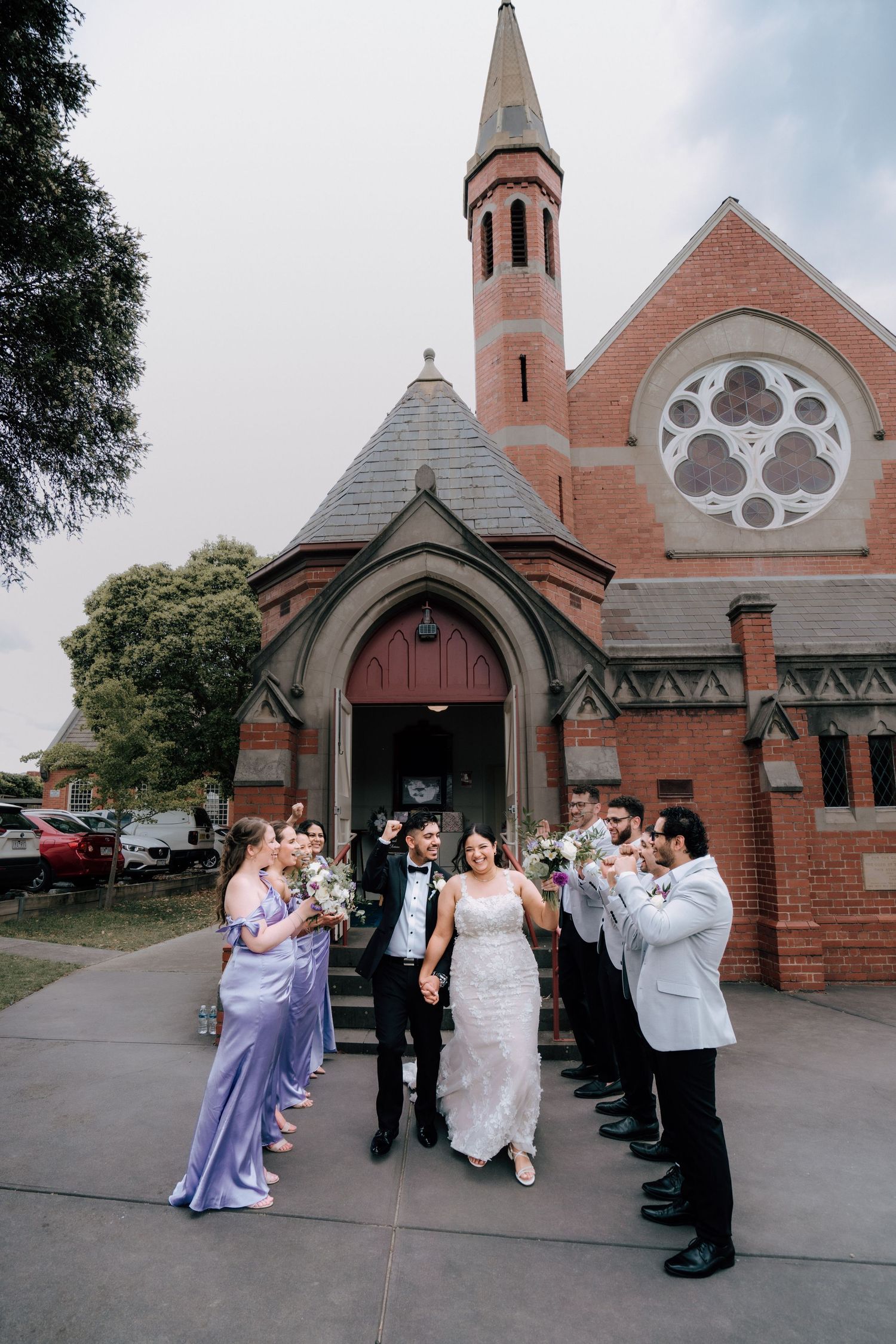 Newlyweds walk hand in hand past cheering wedding party outside red brick heritage church.