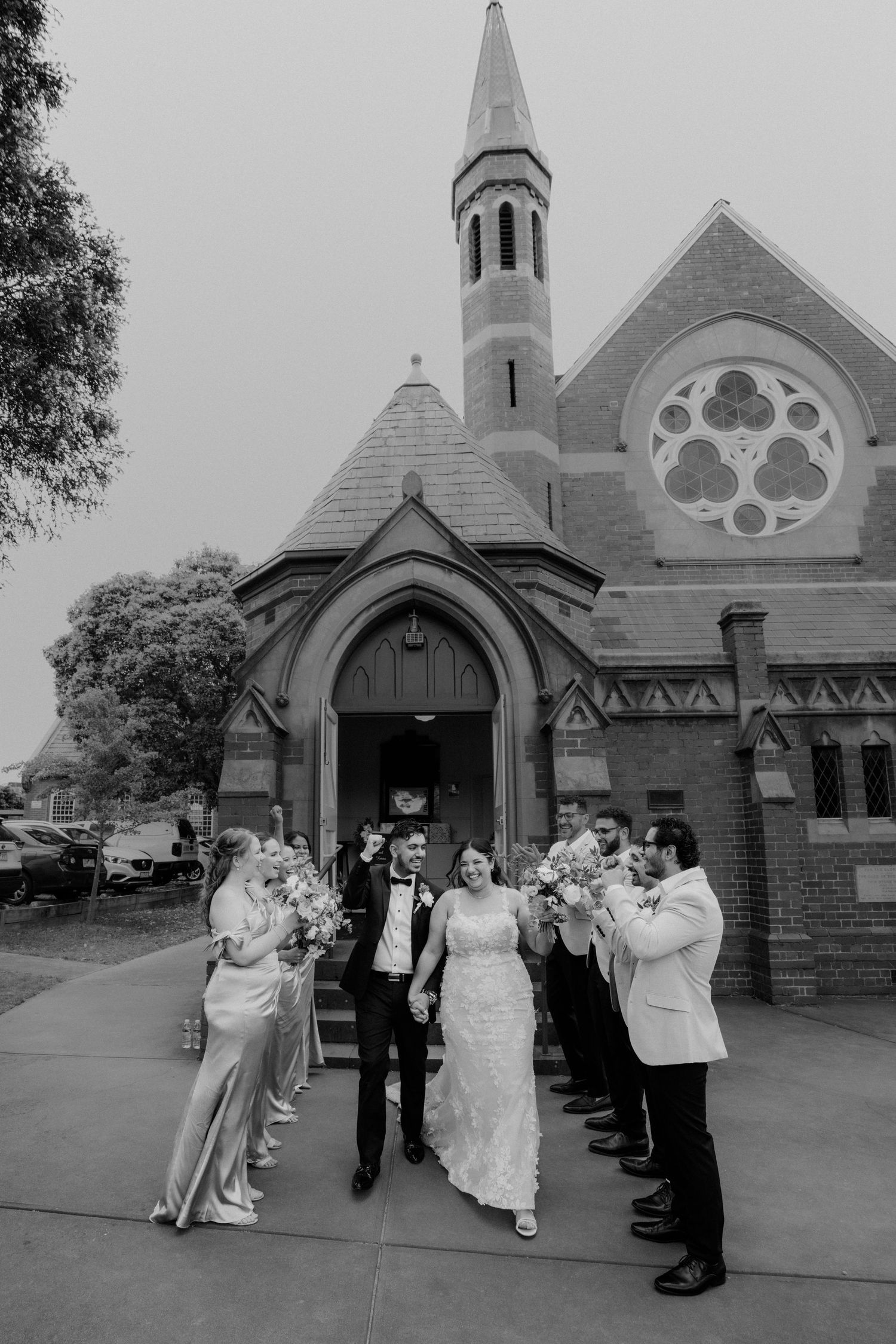 Wedding party exits a historic brick church with rose window, bride and groom leading groomsmen and bridesmaids in black and white.