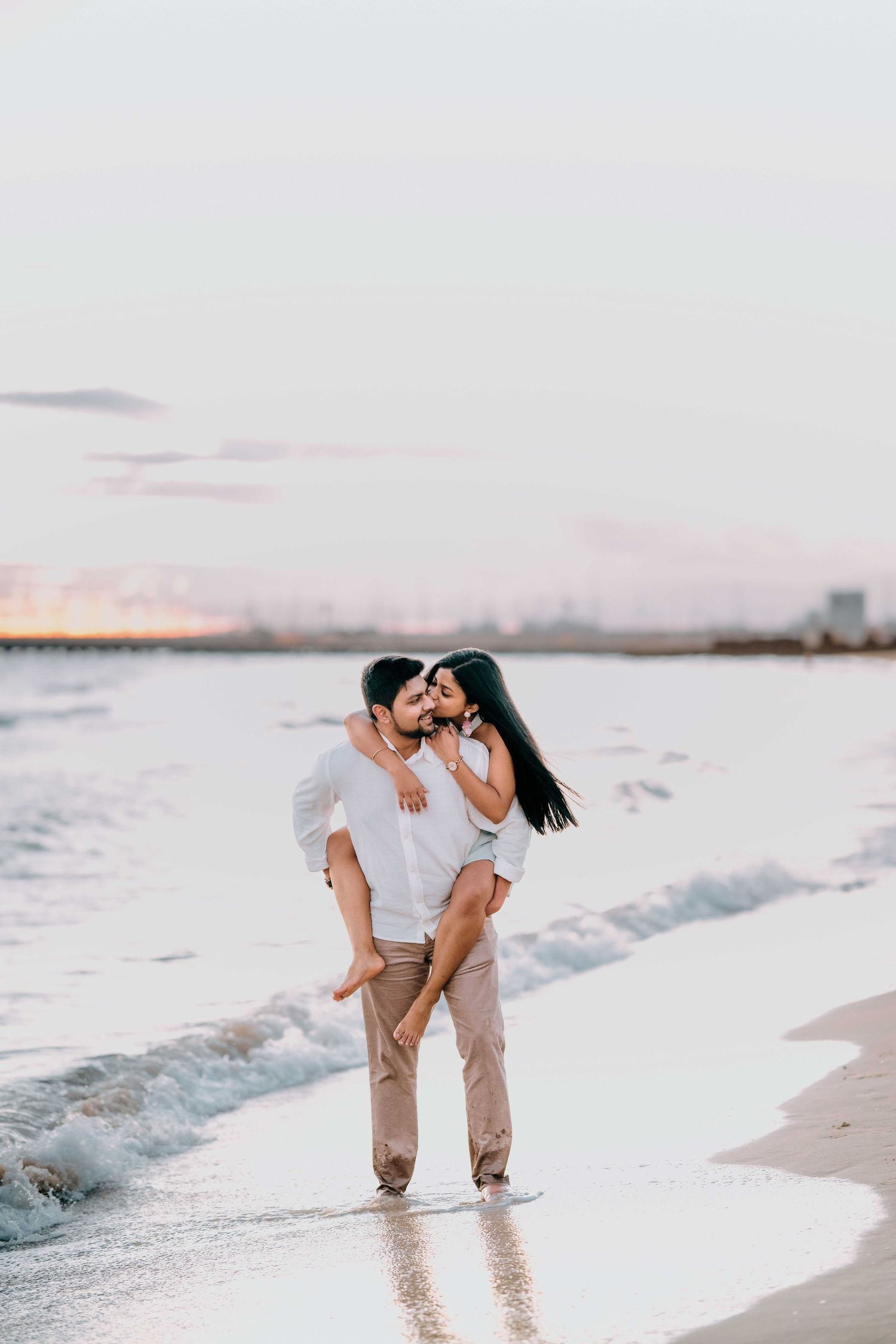 Romantic couple embracing on a beach at sunset, man lifting woman in blue dress by the ocean waves.