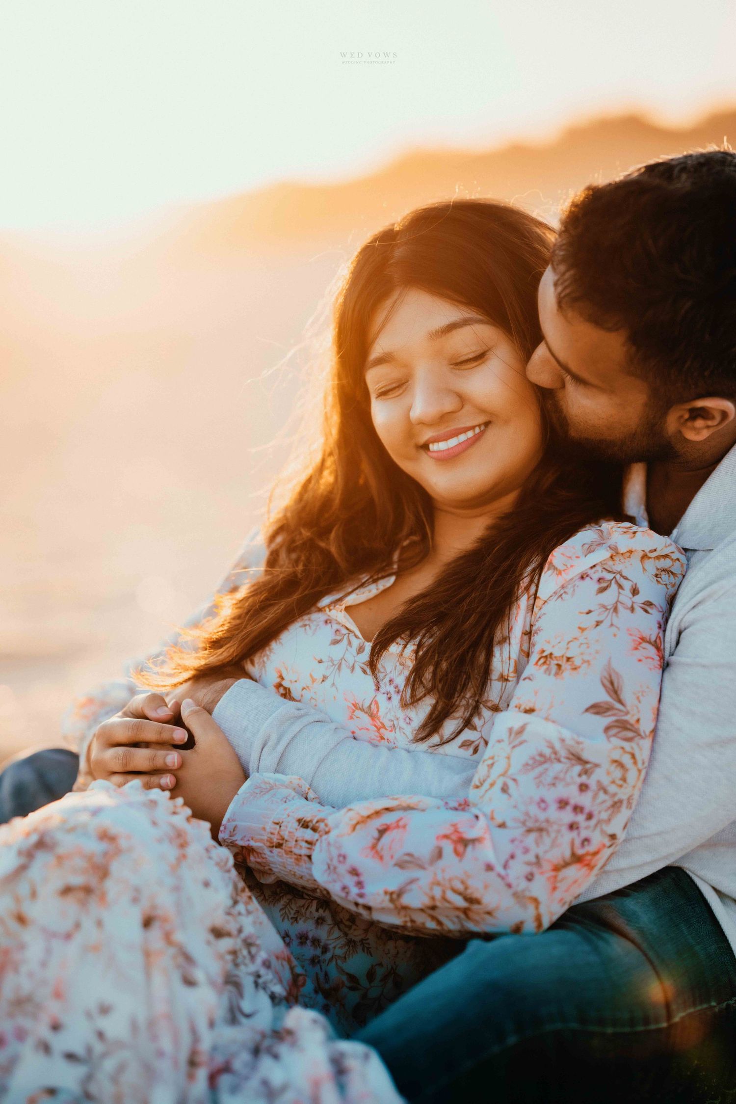Couple sharing tender moment at beach sunset, woman smiling with eyes closed as man embraces her from behind.