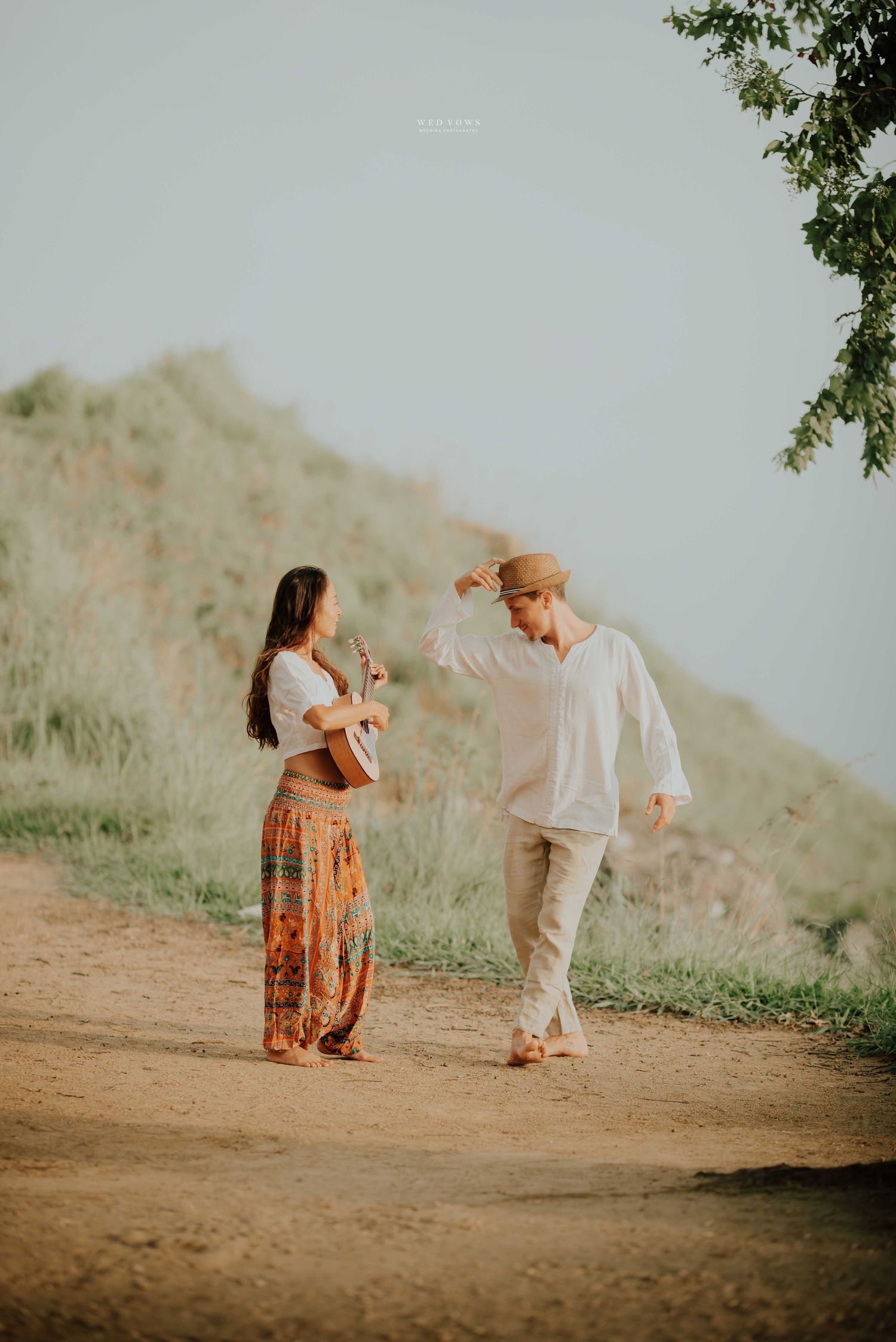 Couple playfully dancing on a dirt path with green hillside in background.