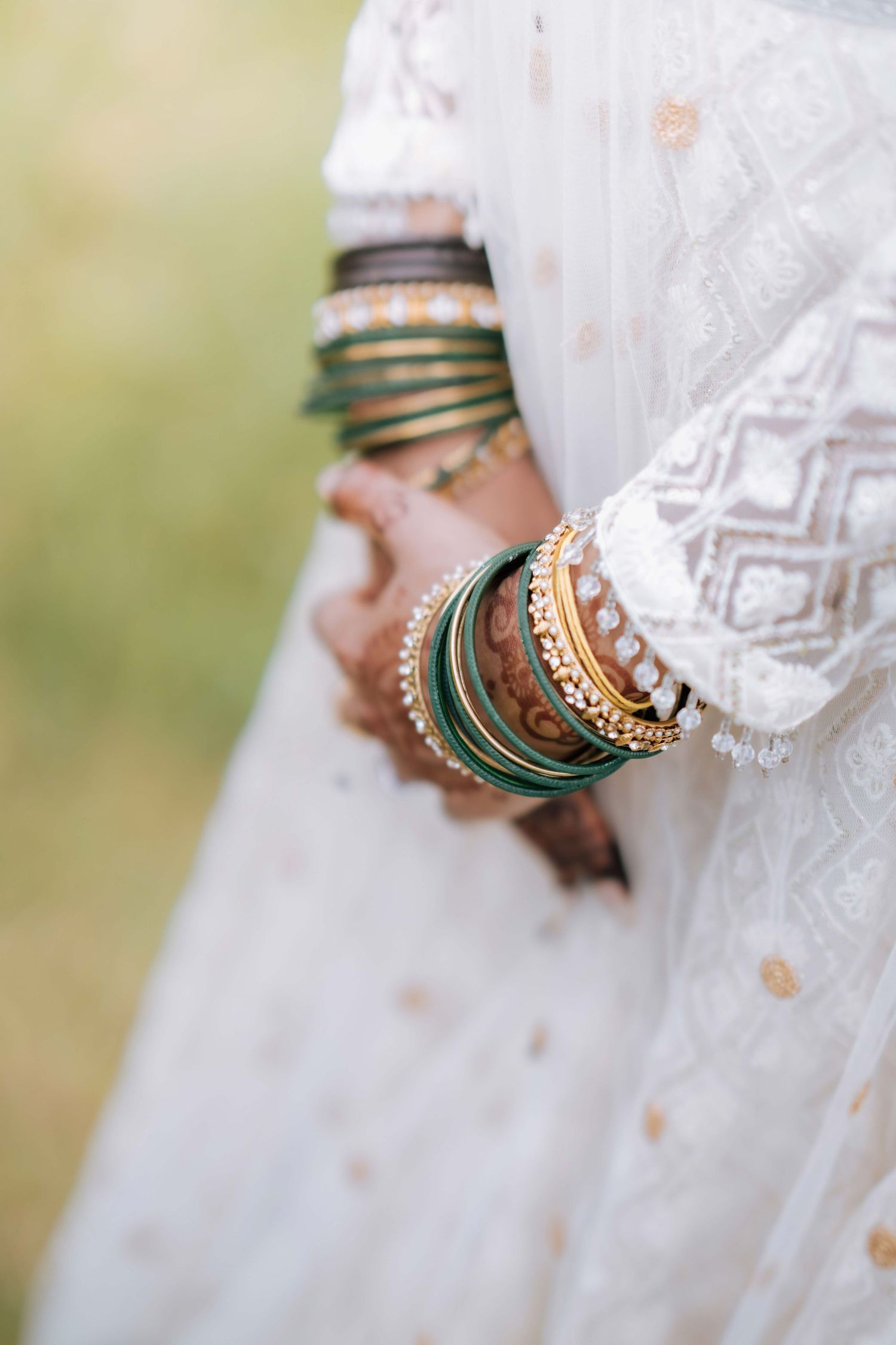 Close-up of Indian bride's wrists adorned with green, gold, and pearl bangles against white lehenga.