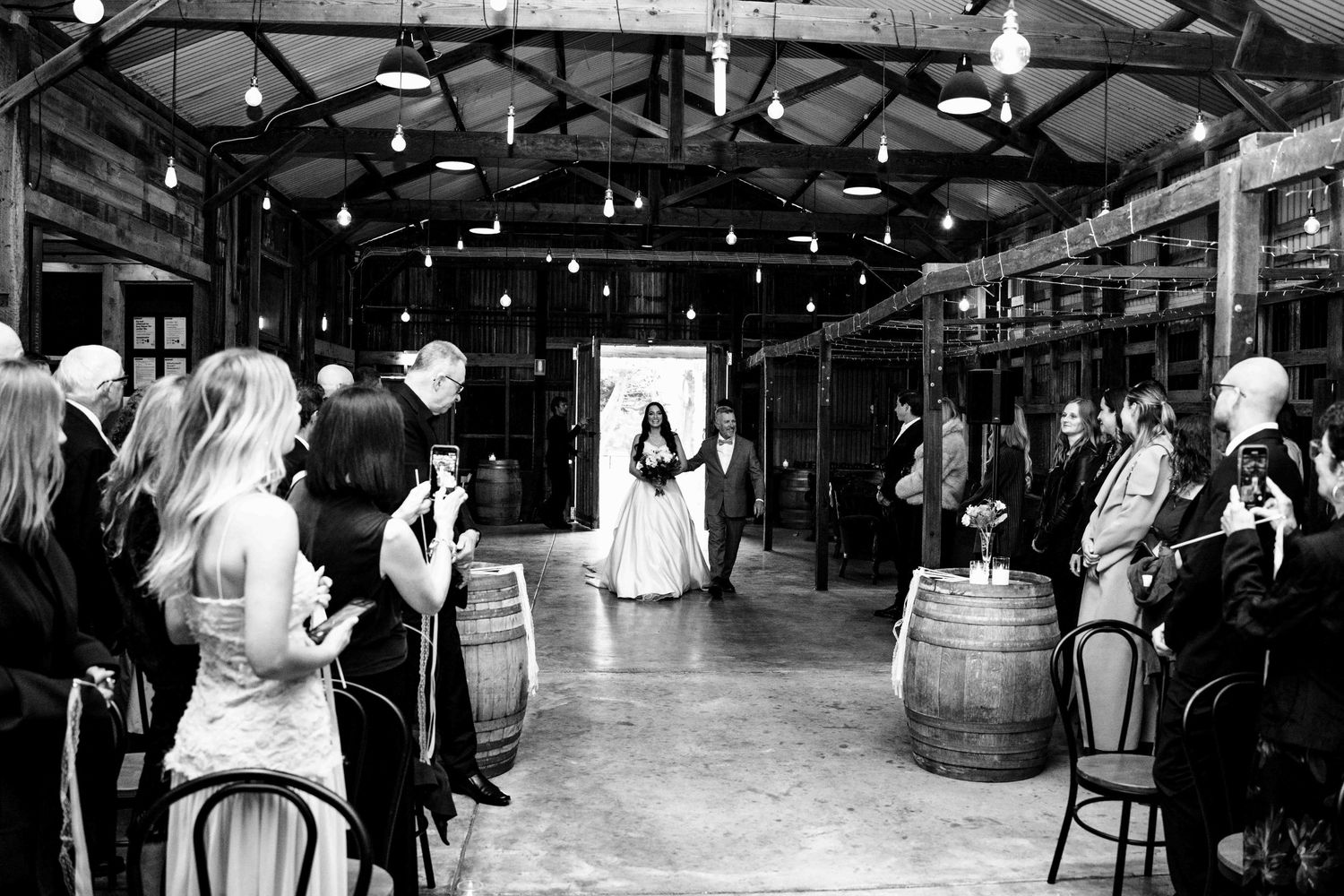 Black and white photo of bride walking down the aisle in a rustic barn wedding ceremony with barrel decorations.