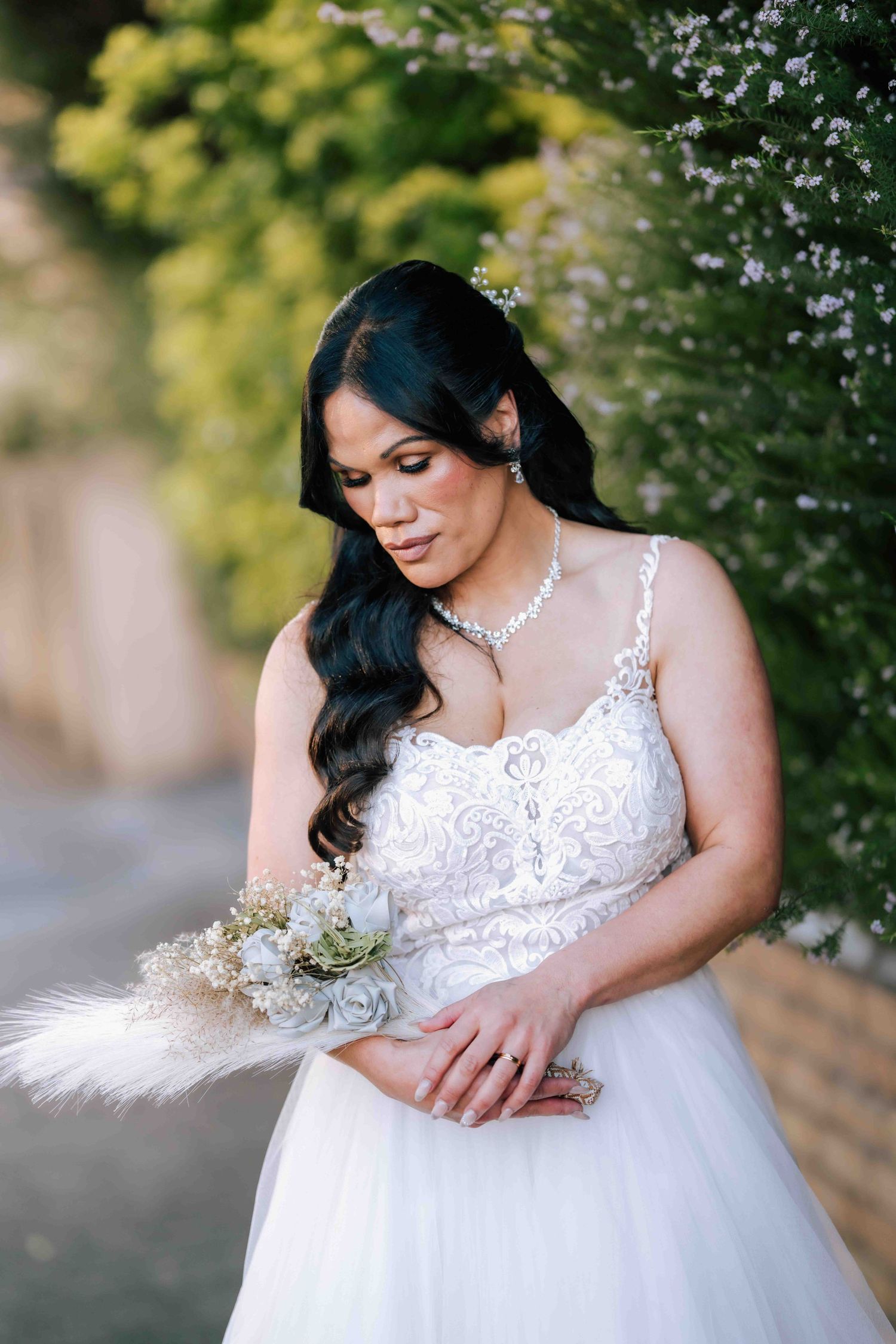 Elegant bride in a white lace gown looking down at her bouquet beside flowering green hedges.