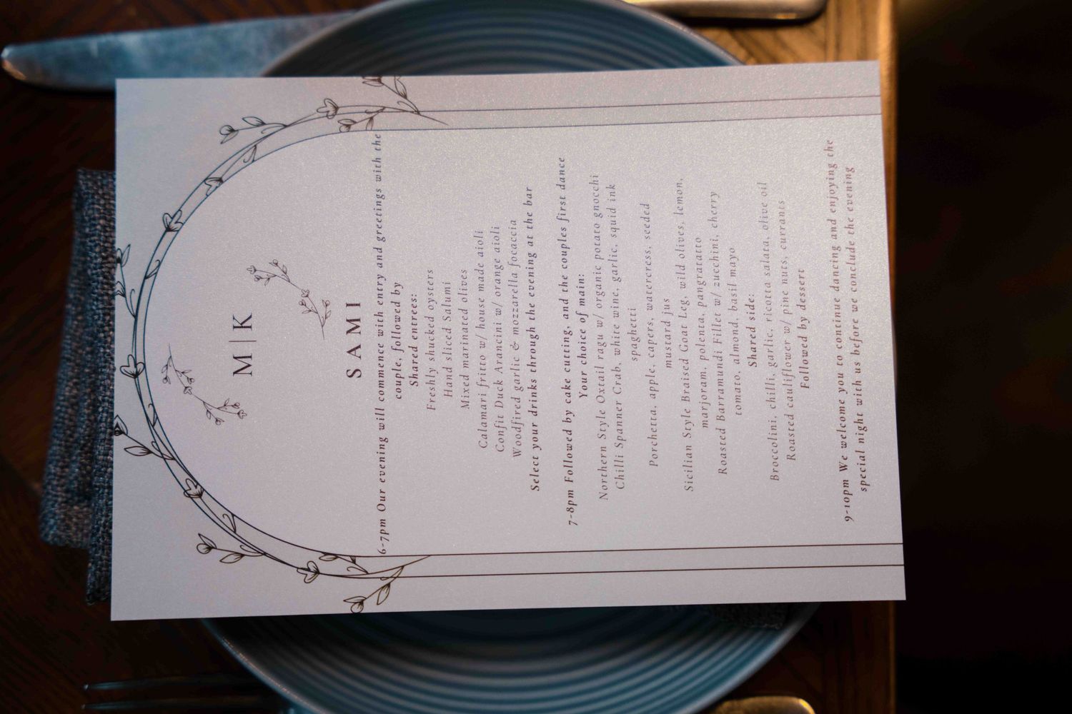 Elegant wedding menu card with botanical wreath design and monogram MK displayed on blue ceramic plate at place setting.
