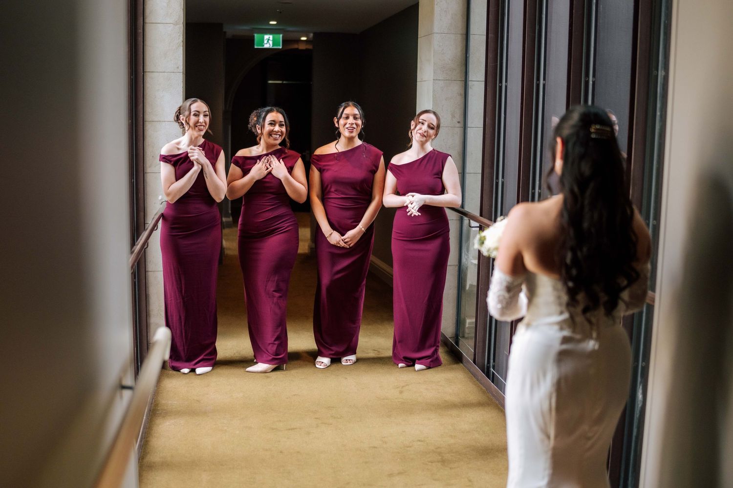 Four bridesmaids in burgundy gowns react with joy as bride in white wedding dress reveals her look.