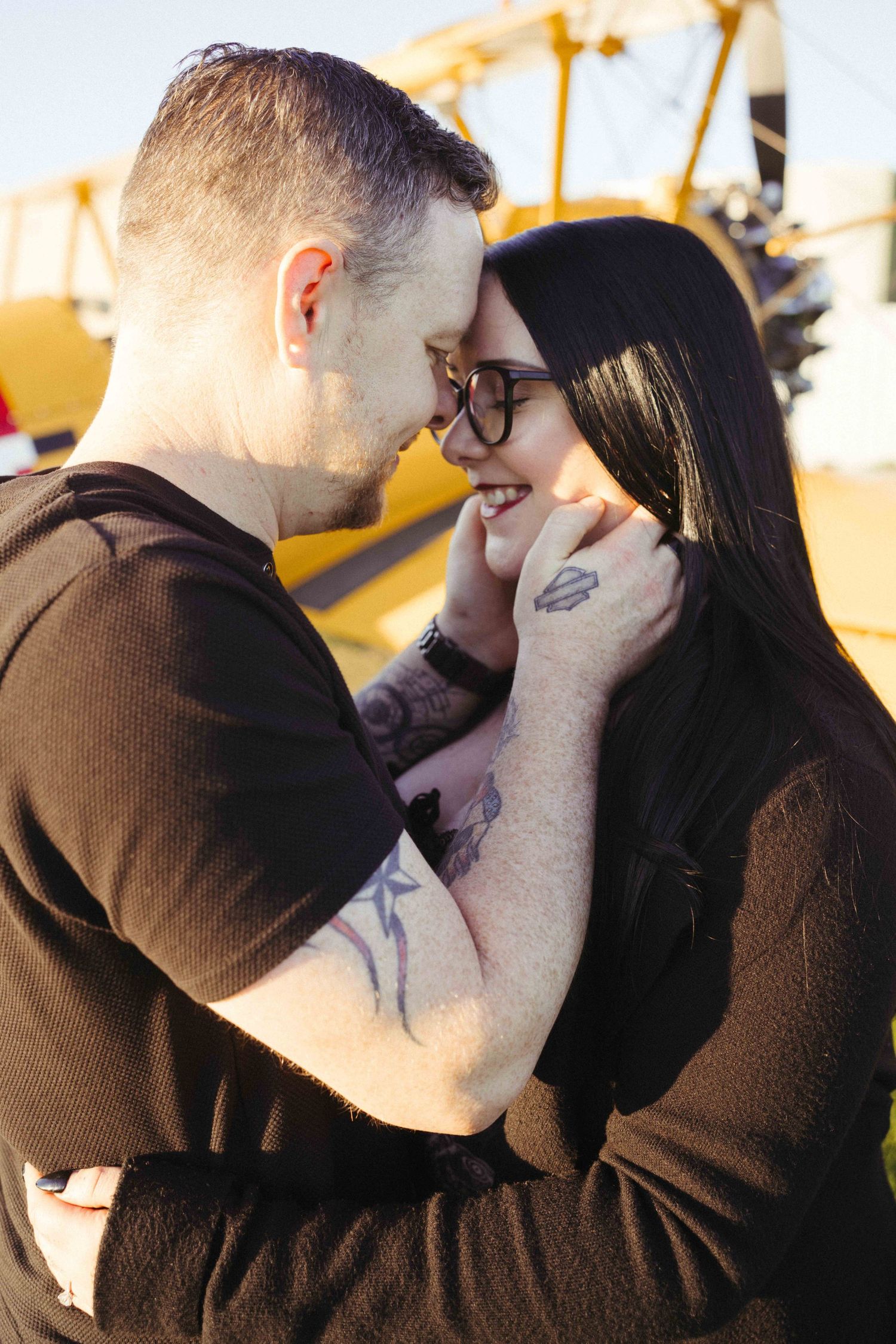 Couple with tattoos sharing a romantic moment, nearly kissing outdoors near a yellow airplane.