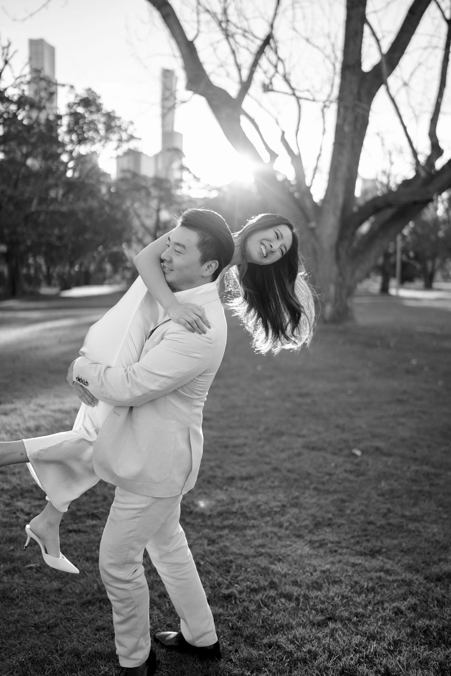 Black and white photo of a man lifting a laughing woman in a park with city buildings in the background.