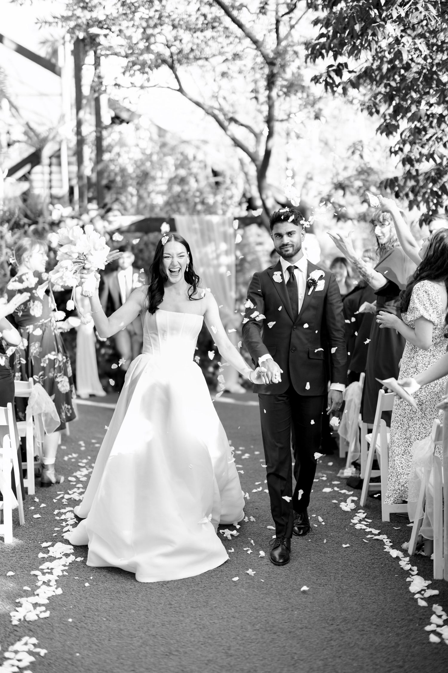 Black and white photo of a joyful bride and groom walking down the aisle as guests toss flower petals.
