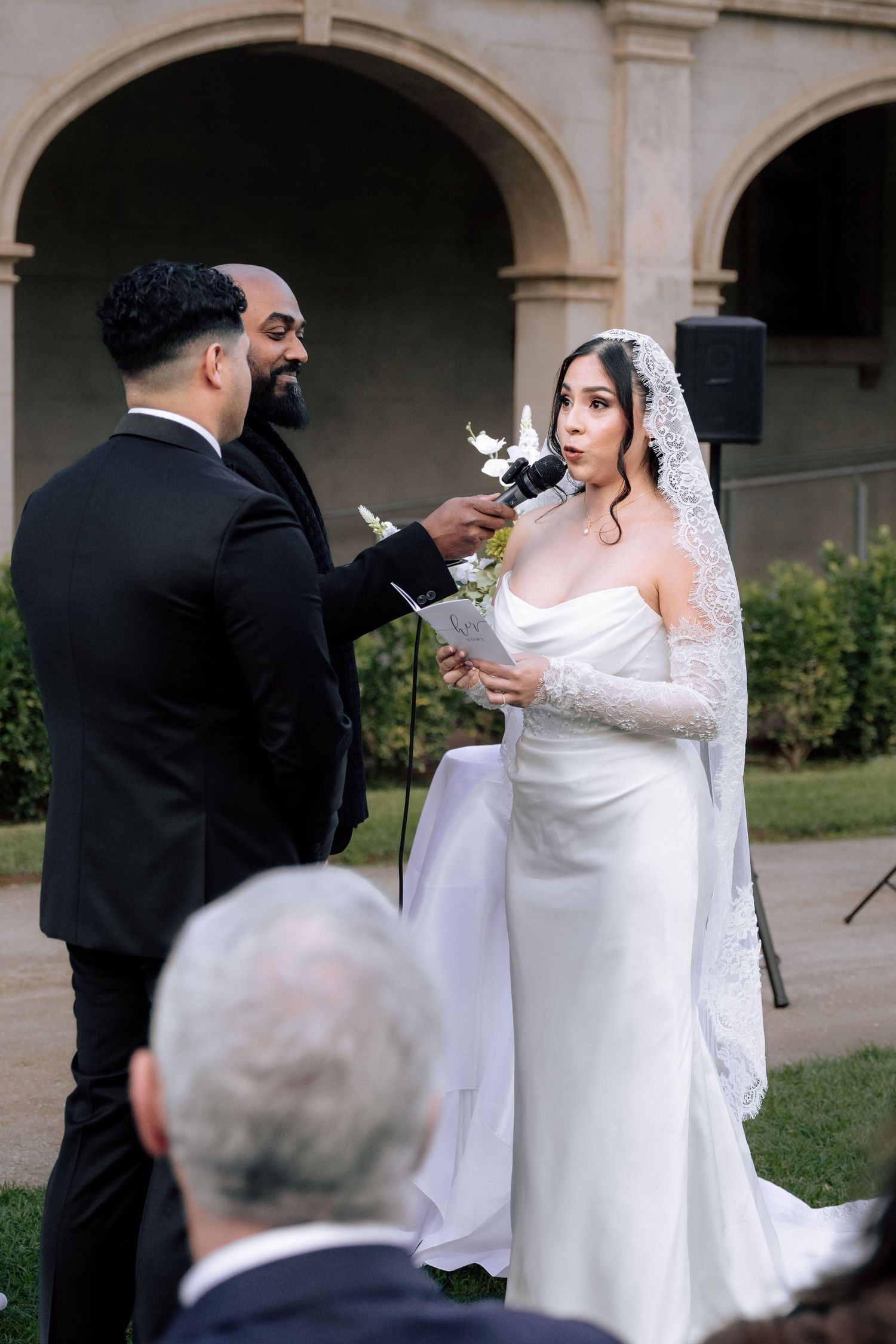 Bride in strapless gown reading wedding vows aloud during outdoor ceremony at historic venue.