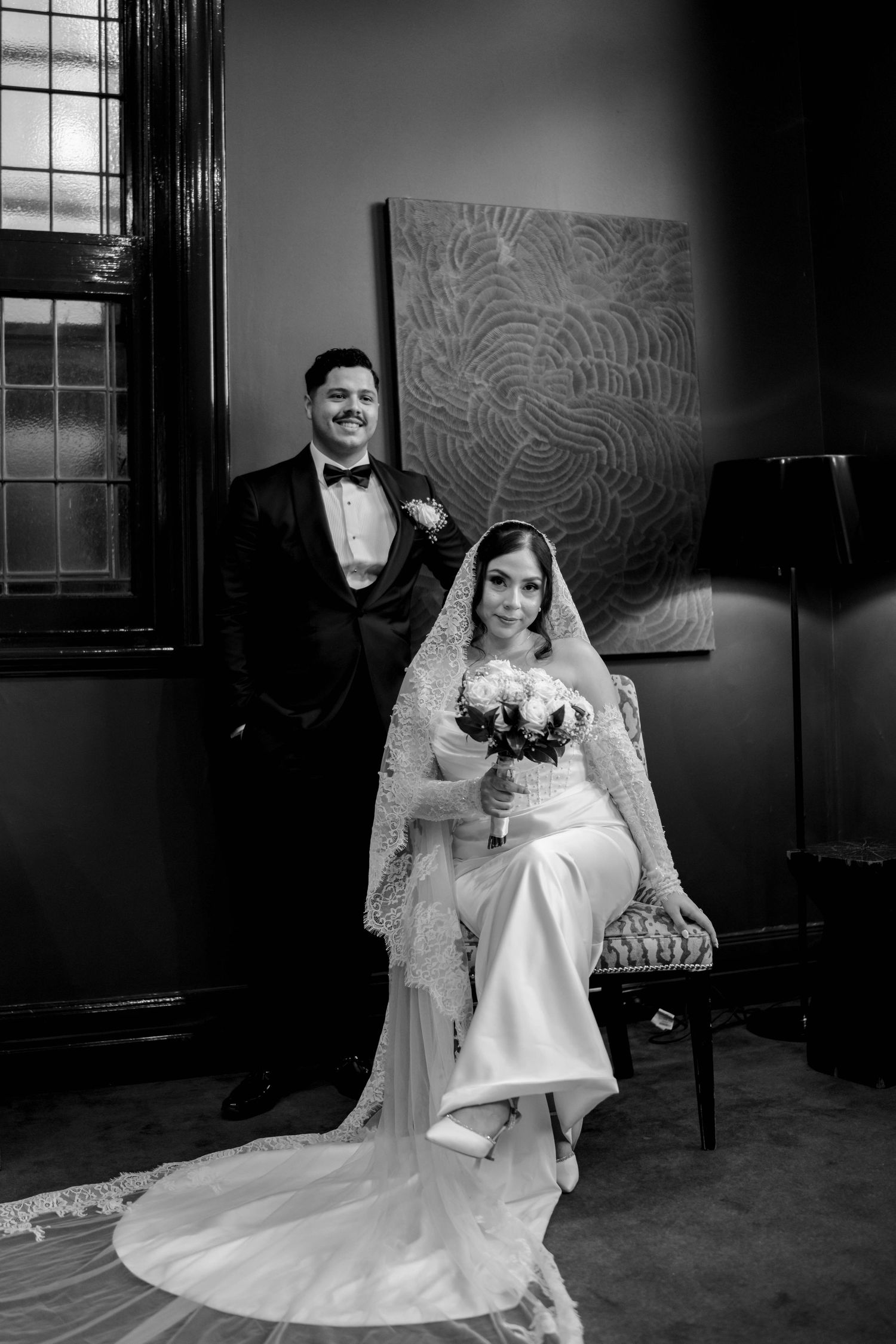 Elegant bride and groom posing in black and white portrait, bride seated in lace veil holding bouquet.