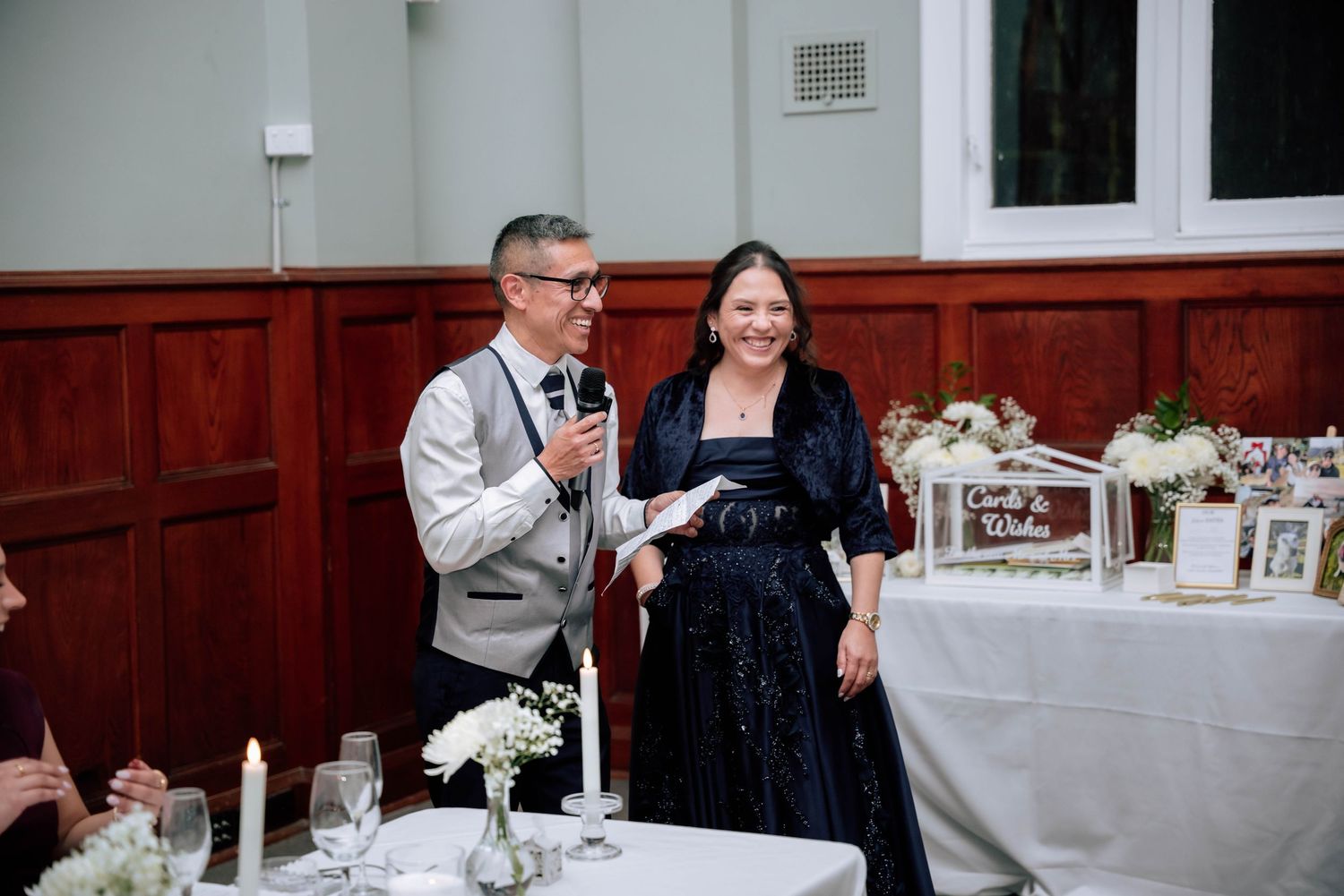 Couple gives speech at wedding reception beside floral arrangements and candlelit tables.