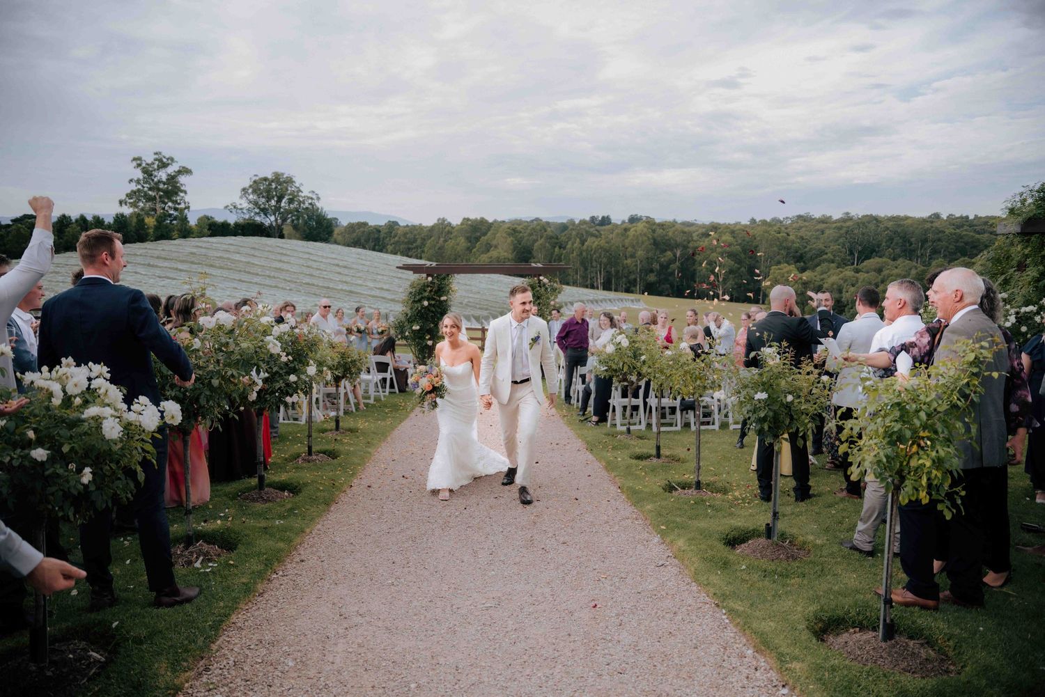 Bride and groom walk down aisle as guests celebrate at outdoor wedding.