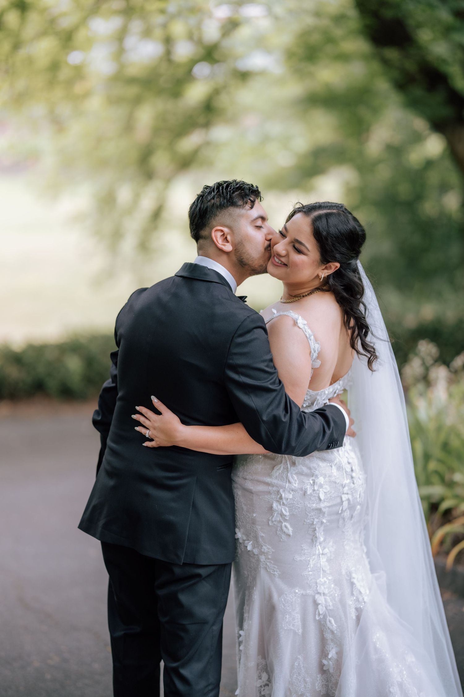 Groom in black suit tenderly kisses bride on cheek during romantic outdoor wedding portrait session.