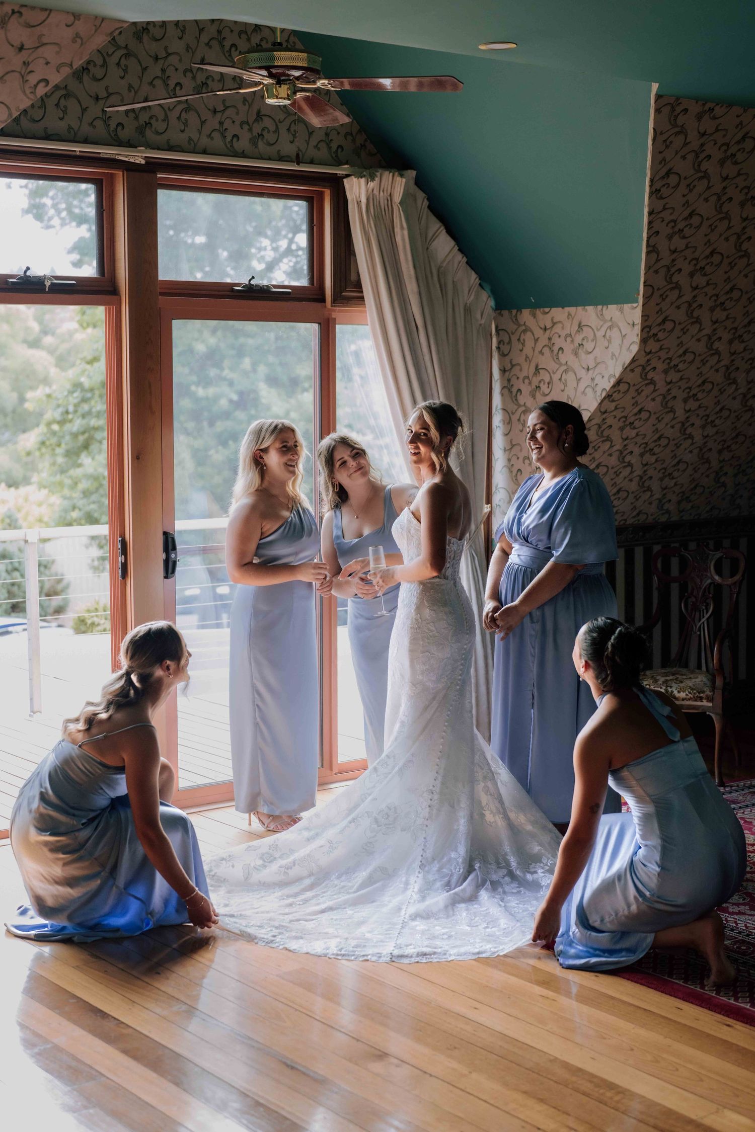 Bridesmaids in blue dresses assist bride with lace gown in sunlit wedding room.