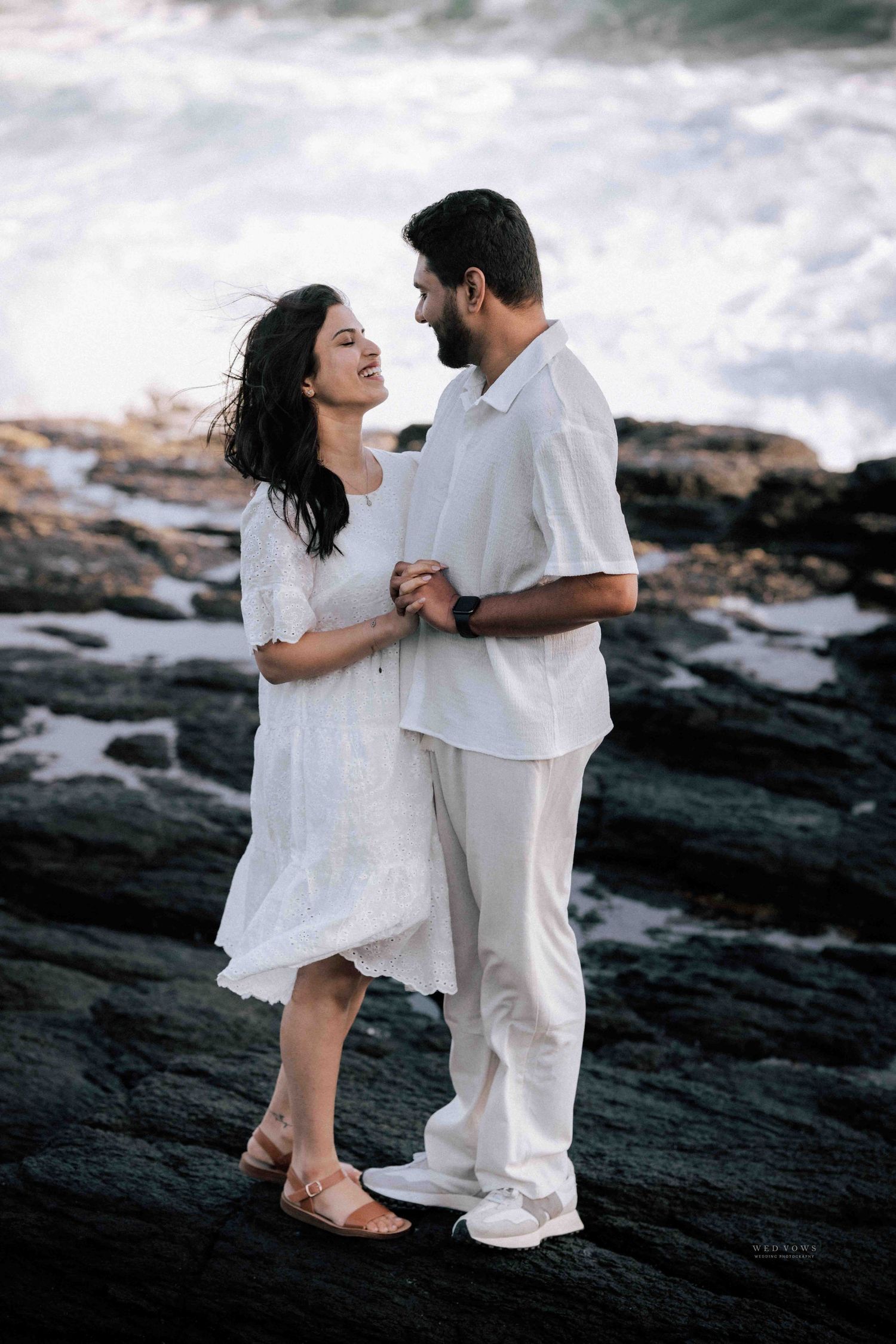 Romantic couple in white outfits sharing a loving gaze on rocky ocean shores.