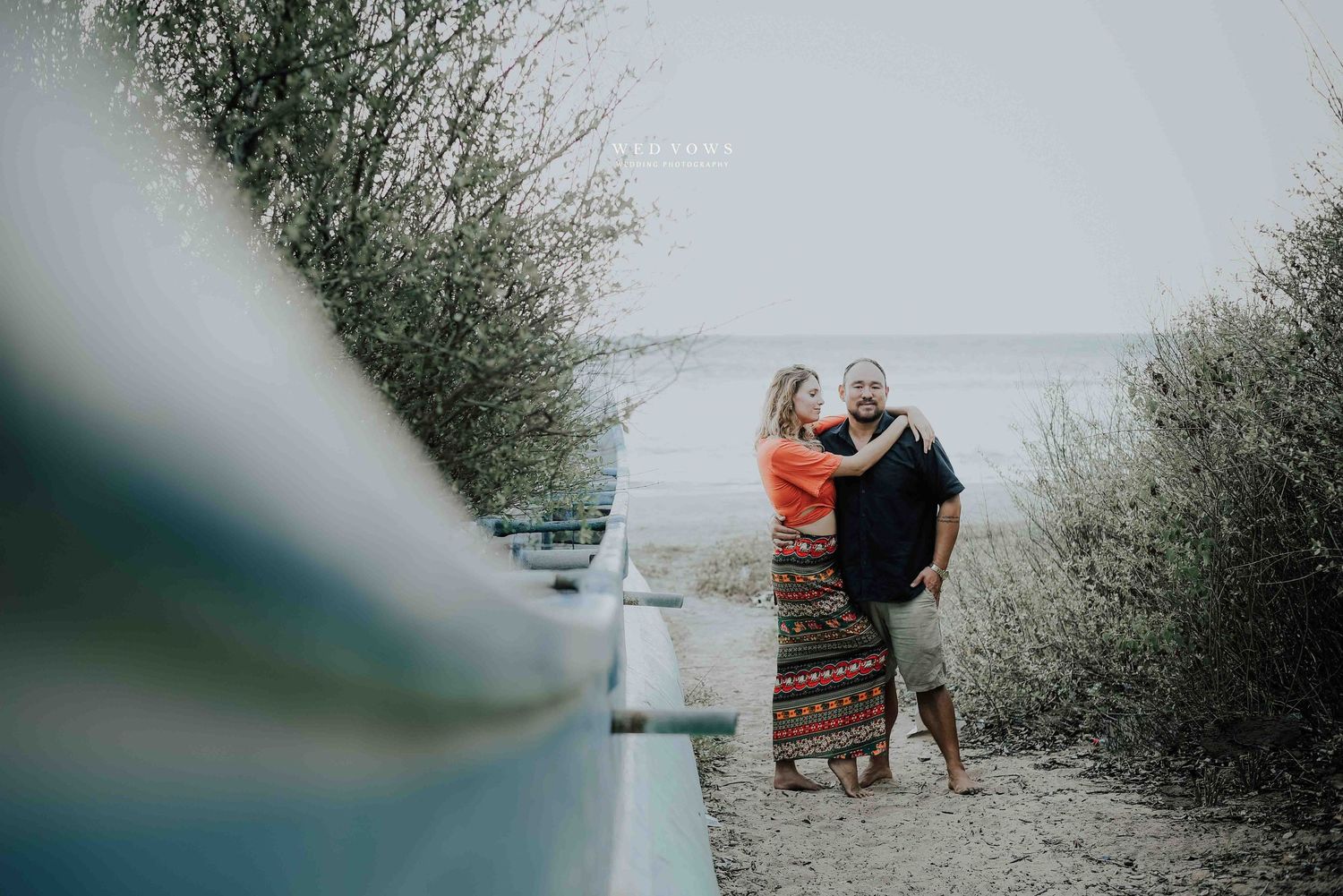 A couple shares a tender embrace on a sandy path between lush bushes with the ocean visible behind them.