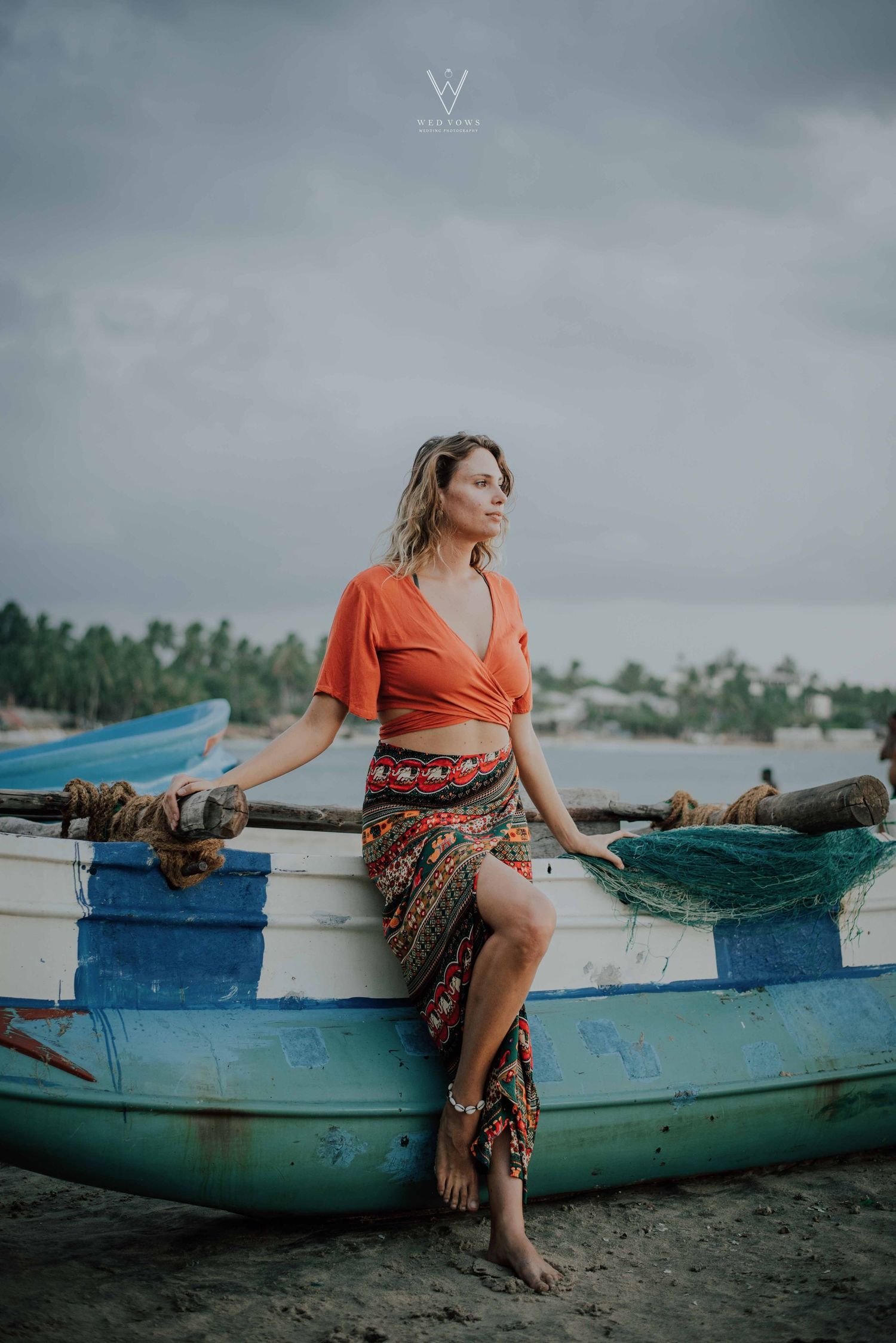 Woman in orange wrap crop top and patterned maxi skirt posing on a rustic blue fishing boat on beach.