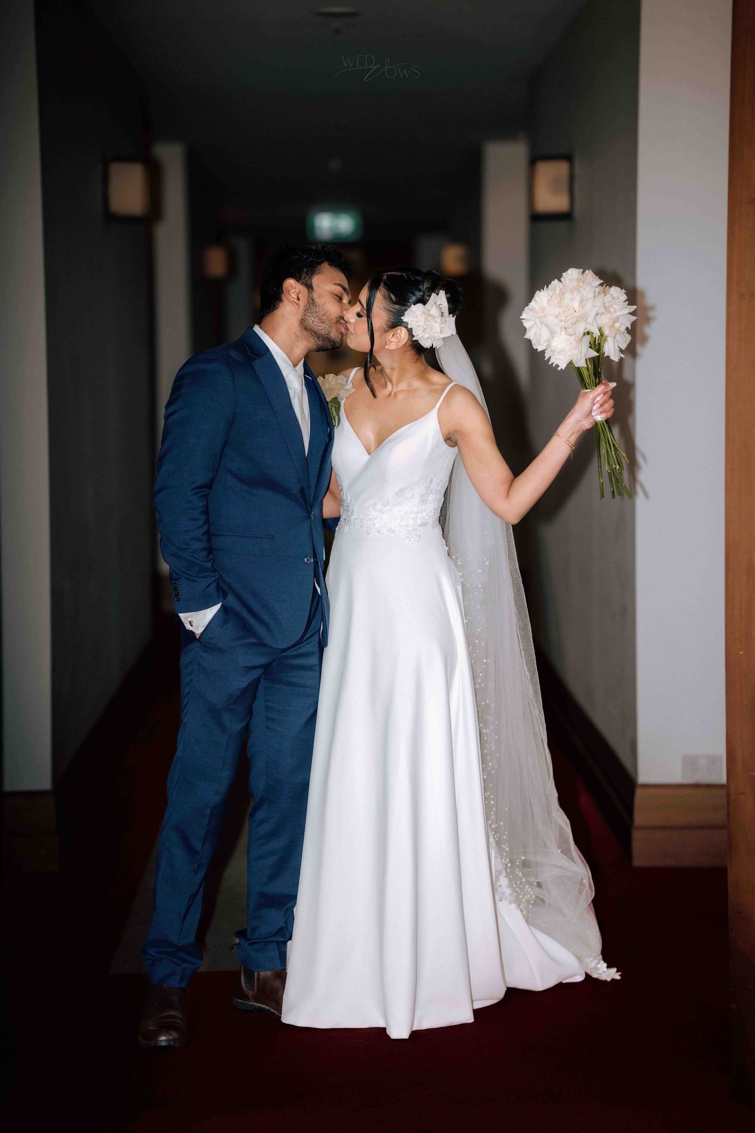 Newlyweds sharing a kiss in a hotel hallway, groom in navy suit, bride holding white bouquet.
