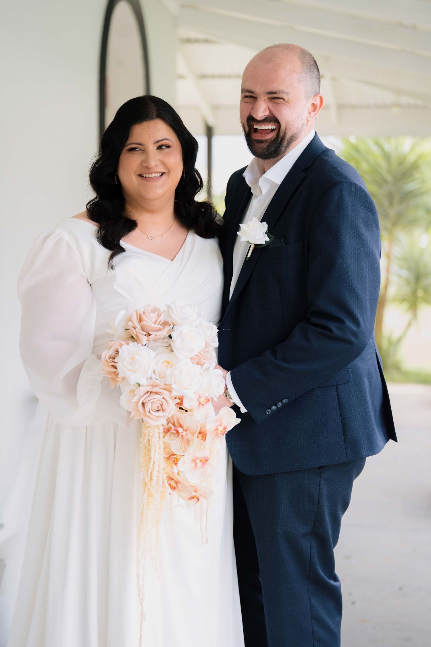Smiling bride and groom posing together, bride holding cascading blush and white floral bouquet in white wedding dress.