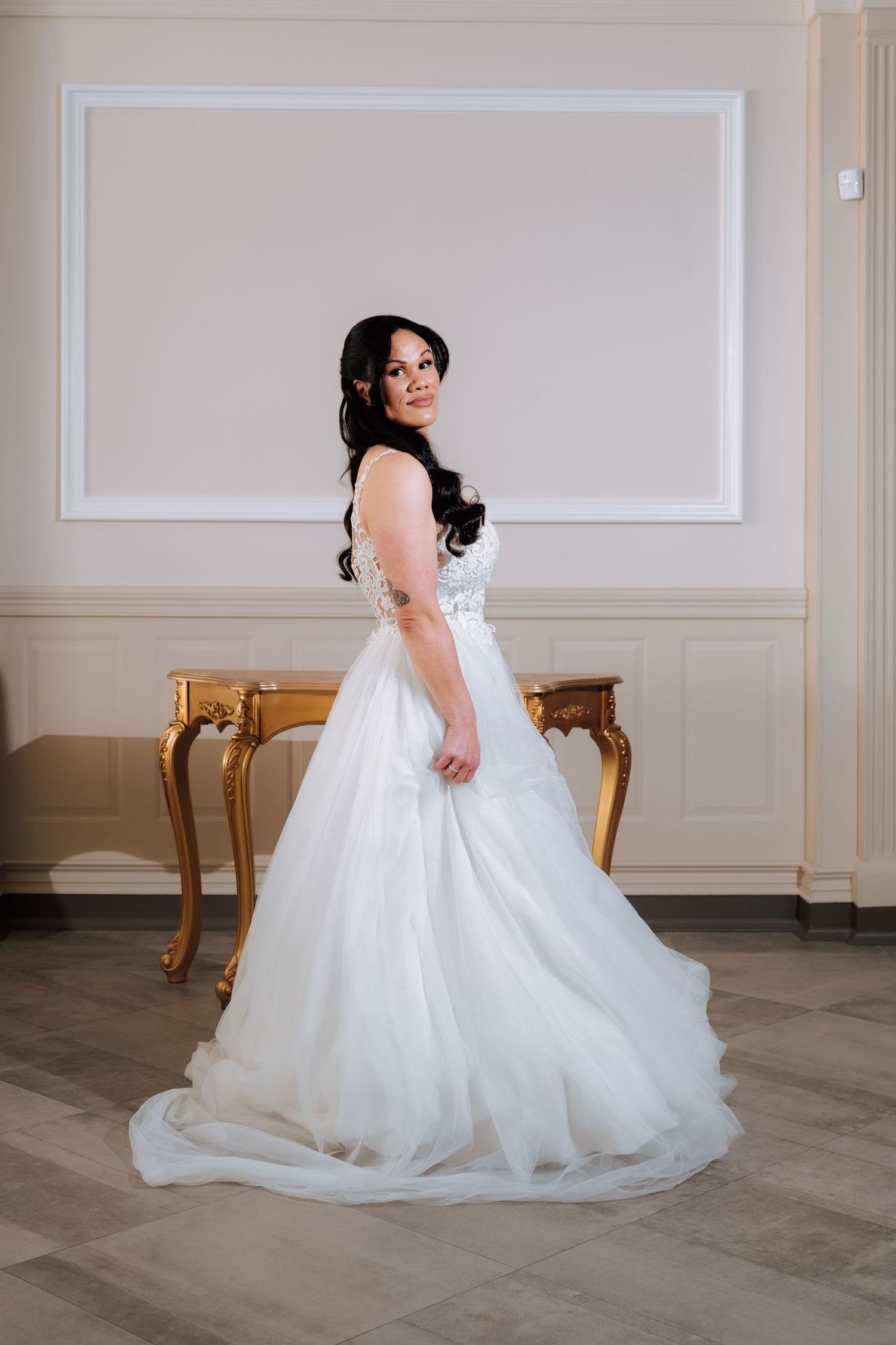 Elegant bride in a white ball gown standing in a classic interior with a gold ornate table.