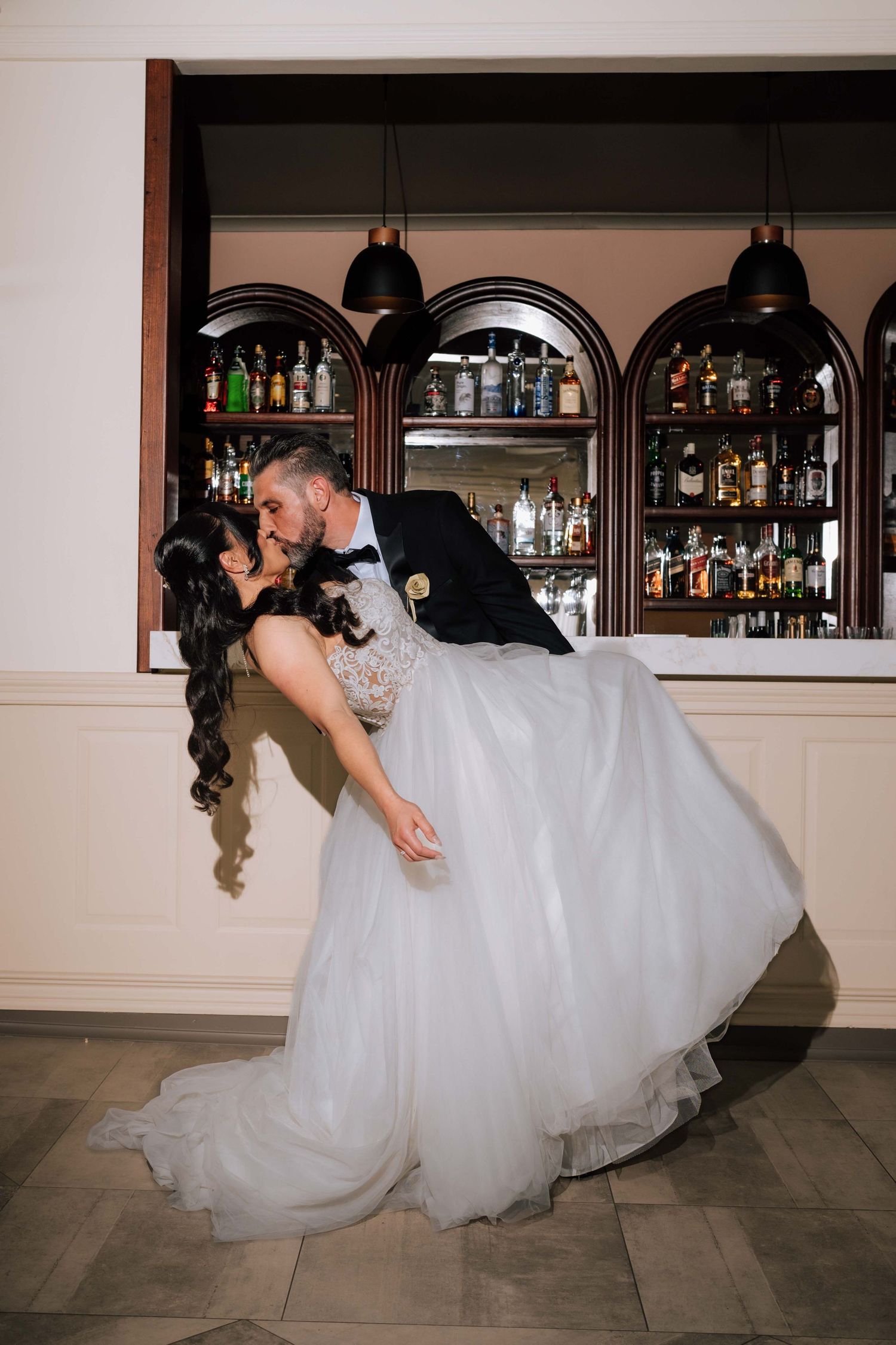 Groom dipping bride in a romantic kiss in front of a bar with arched shelves of bottles behind them.