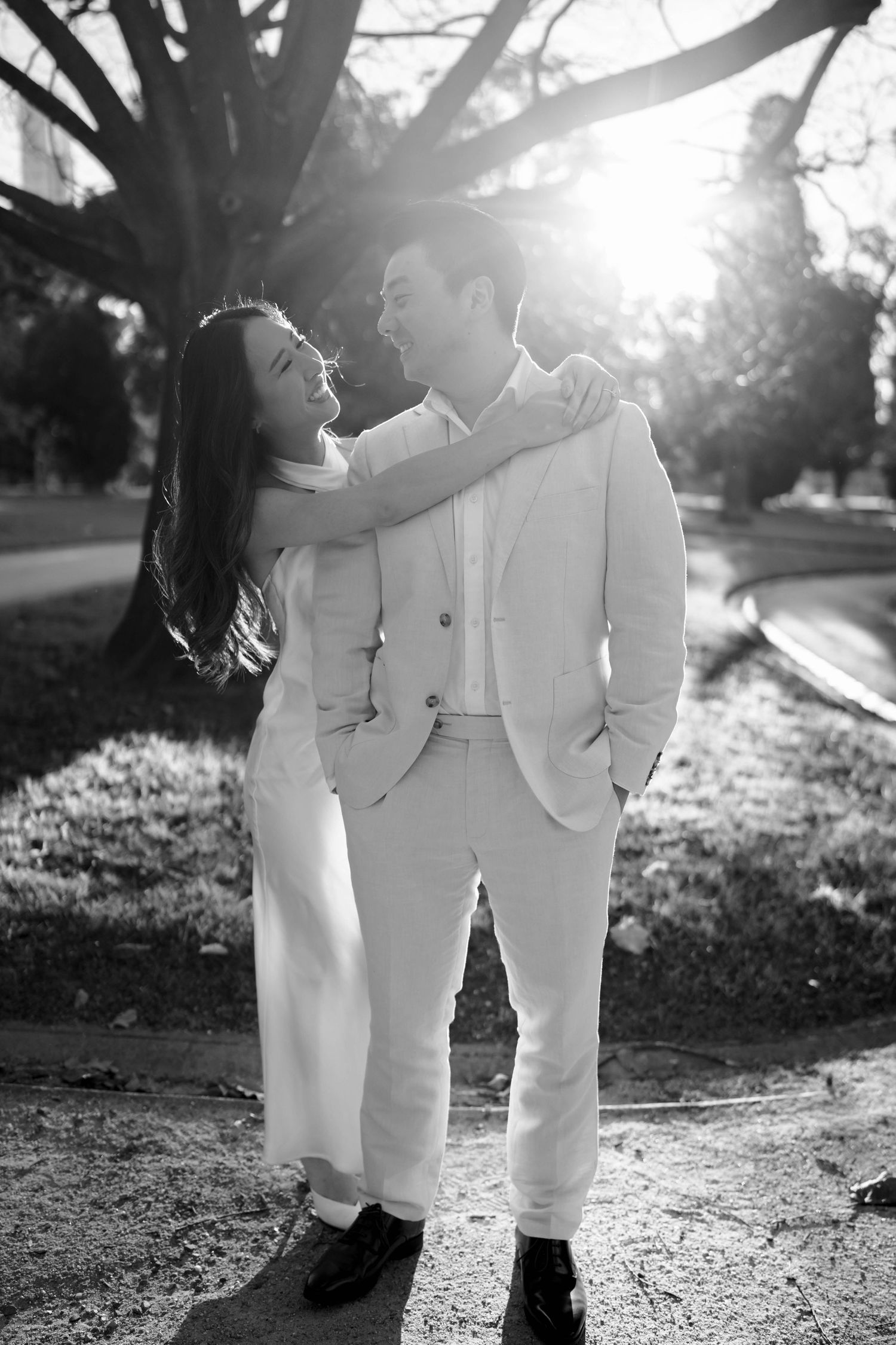 Black and white portrait of Asian couple embracing and smiling at each other in a sunlit park setting.