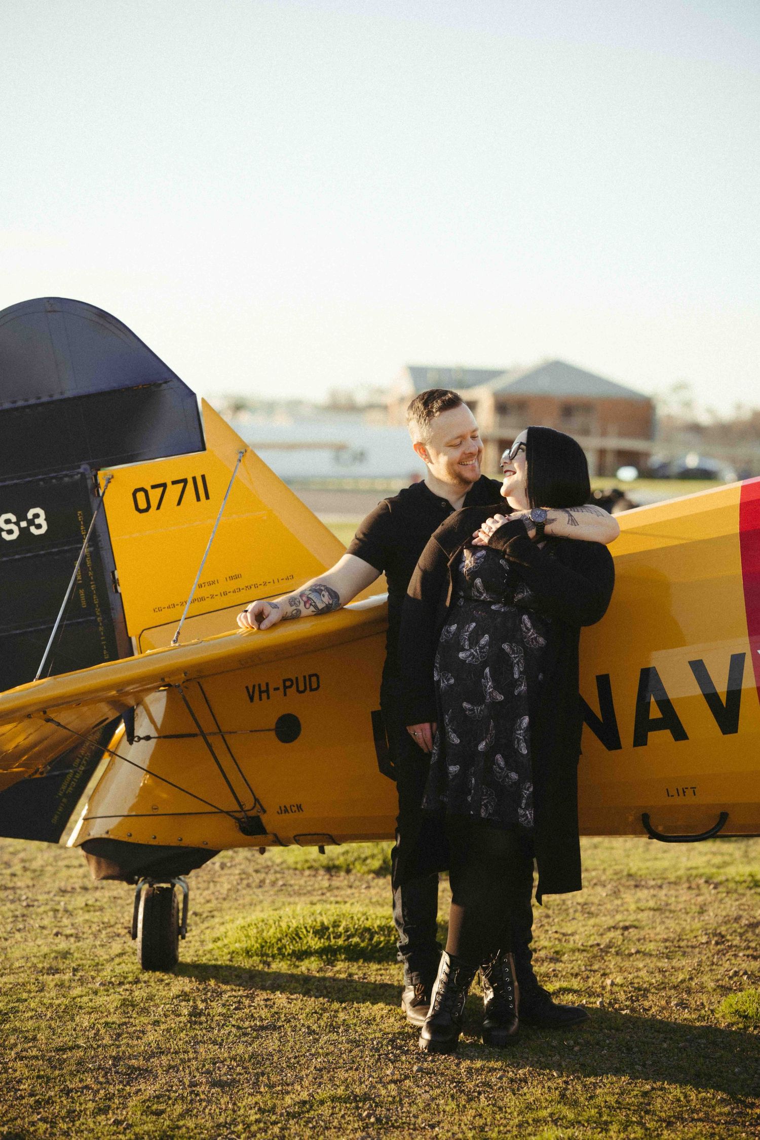 Couple embracing romantically beside a vintage yellow Navy aircraft numbered 07711 on a sunny airfield.