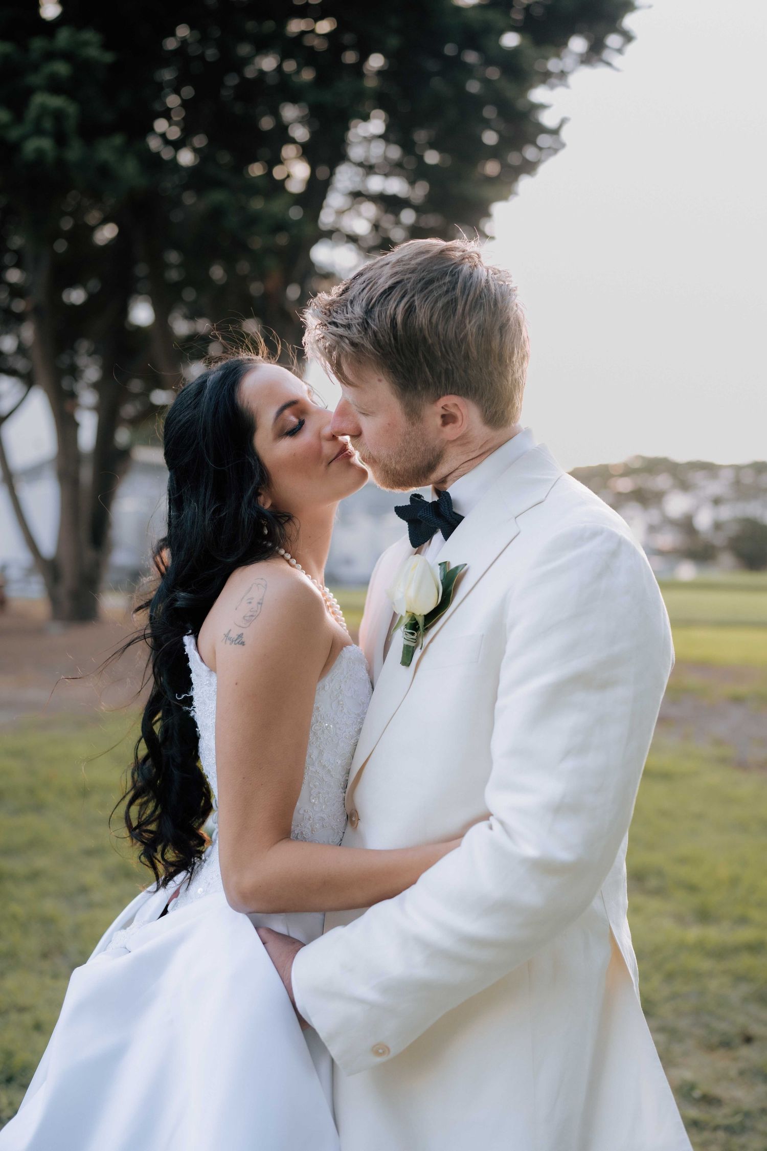 A bride and groom share a romantic kiss outdoors, with the groom in a white tuxedo and the bride in a white wedding gown.