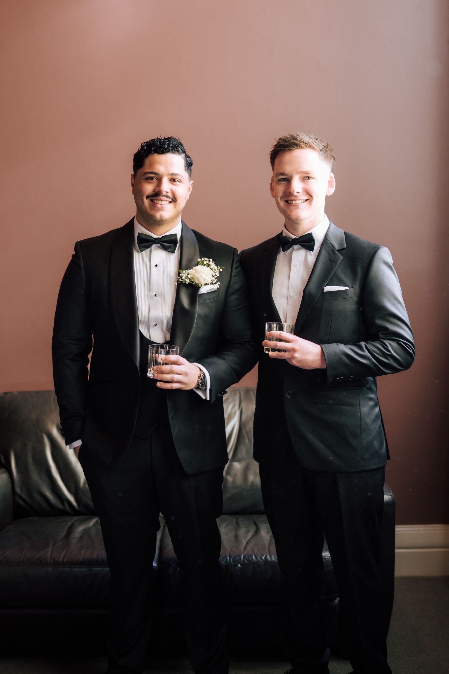 Two grooms in black tuxedos with bow ties holding champagne glasses at their wedding celebration.
