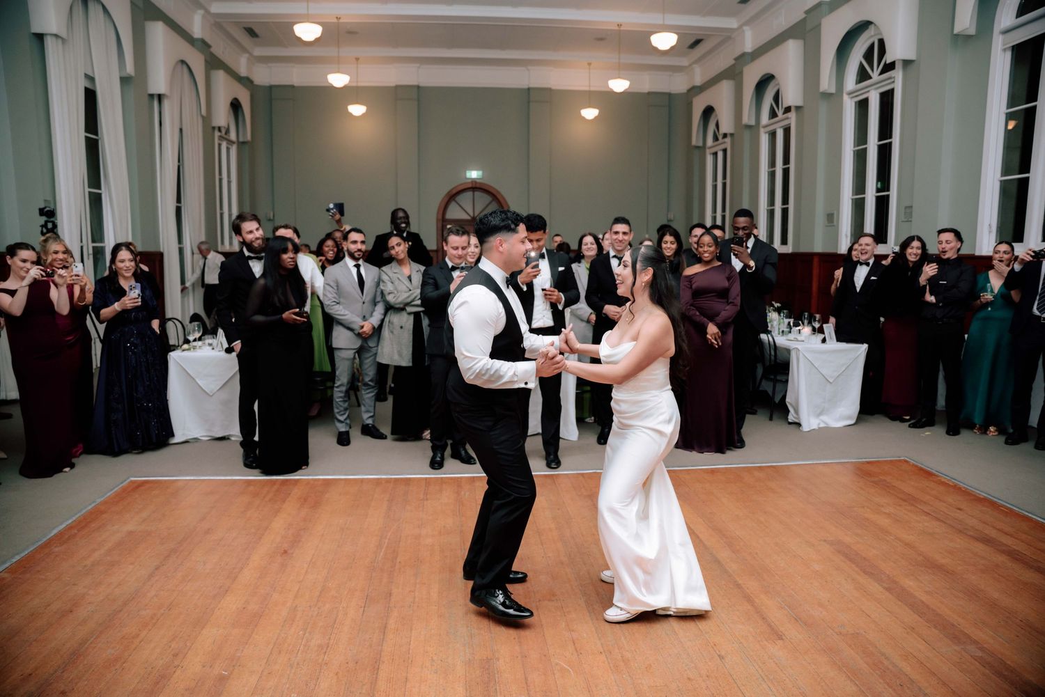 Bride and groom share first dance on wooden dance floor surrounded by wedding guests.