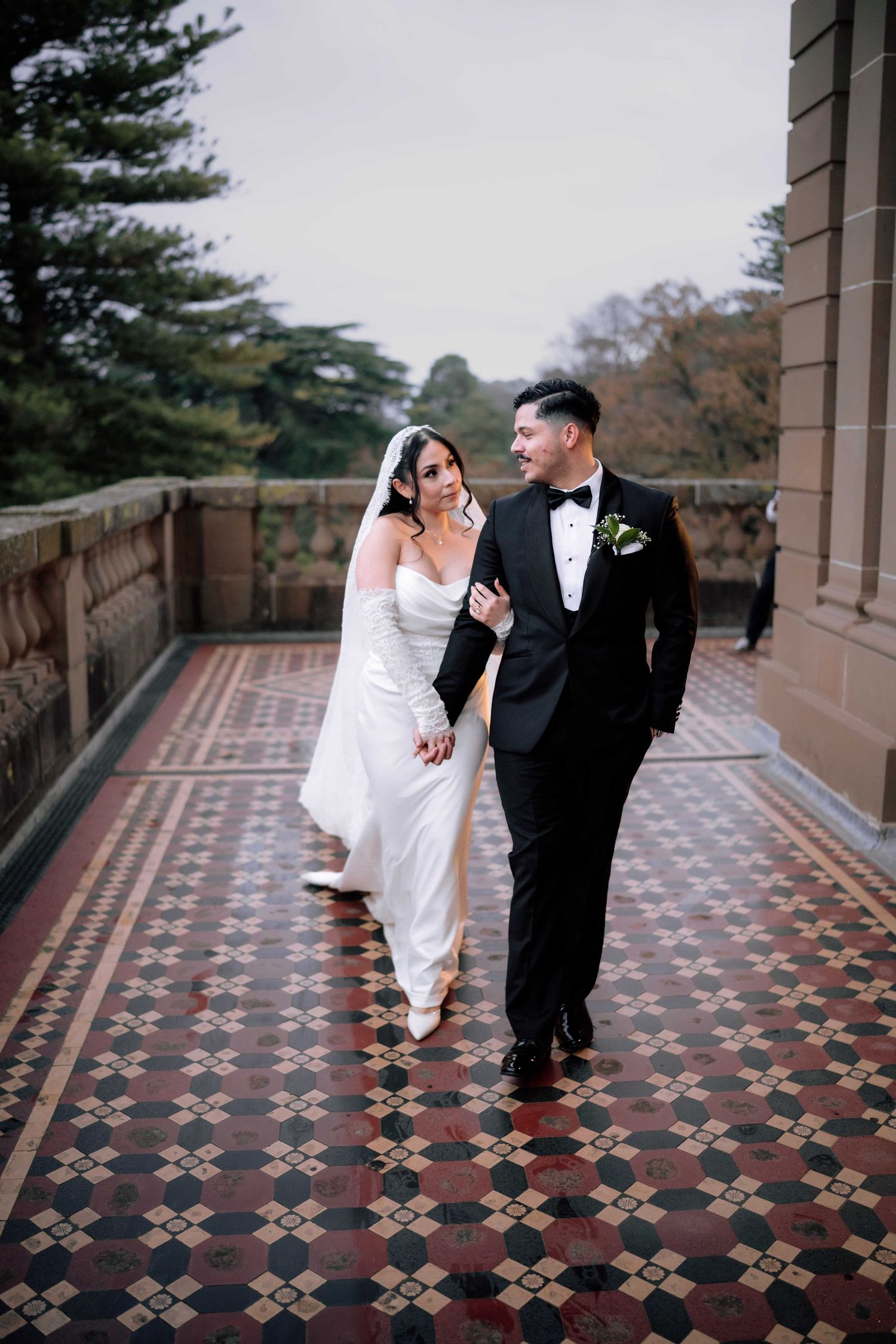 Bride in sleek white gown and groom in black tuxedo walk together on ornate tiled terrace.