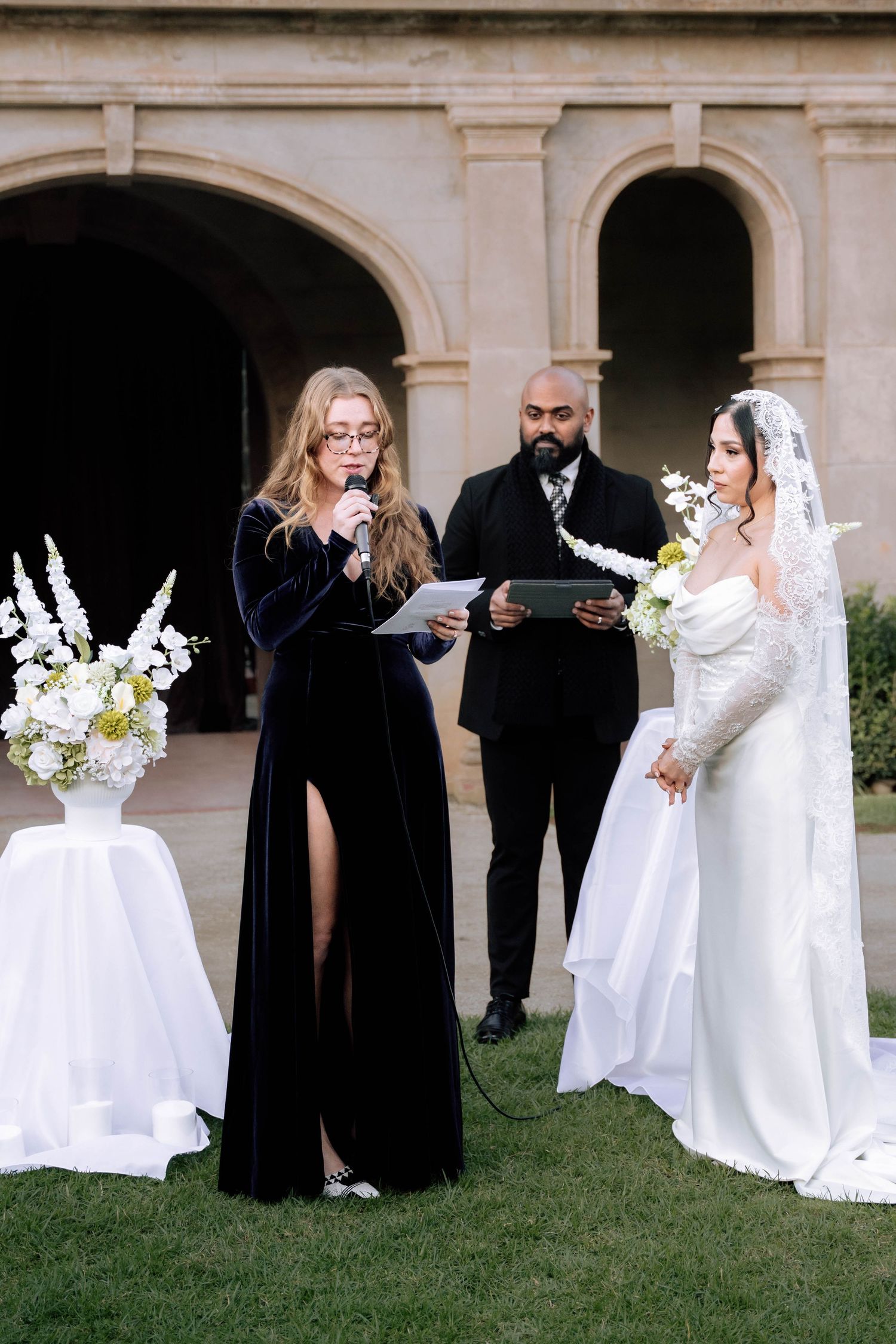 Woman in navy velvet gown reads speech at outdoor wedding ceremony beside bride in white lace gown and veil.