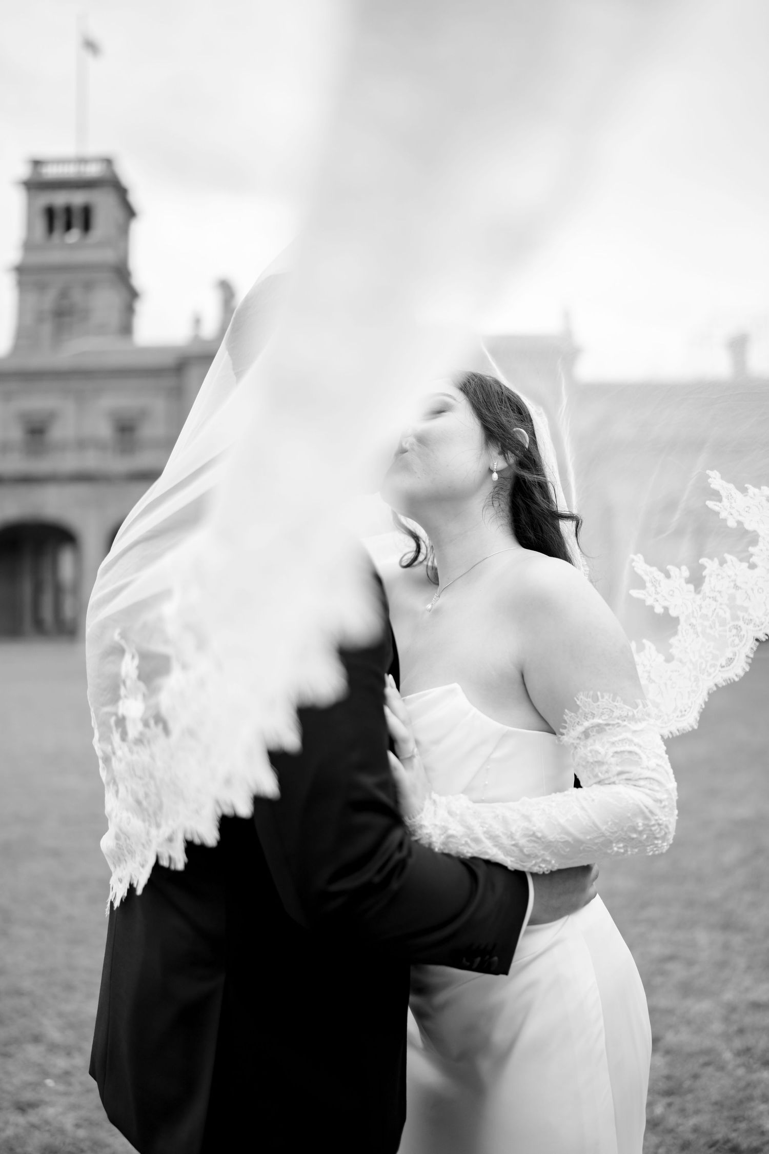 Black and white photo of bride and groom embracing as lace veil billows in wind at historic venue.