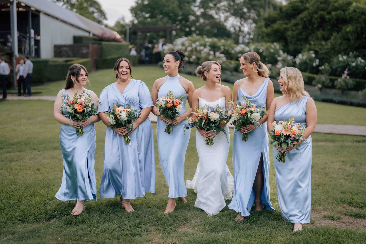Bride and five bridesmaids in light blue satin gowns walking together holding colorful bouquets.