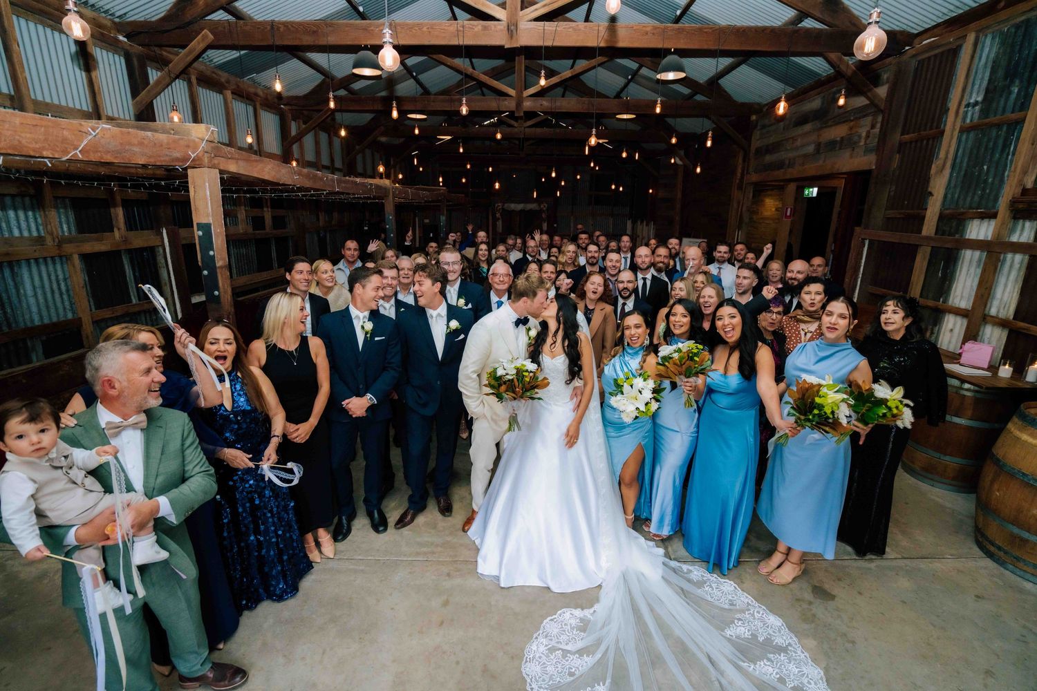 Wedding couple kisses surrounded by guests and bridesmaids in blue dresses inside a rustic barn venue.