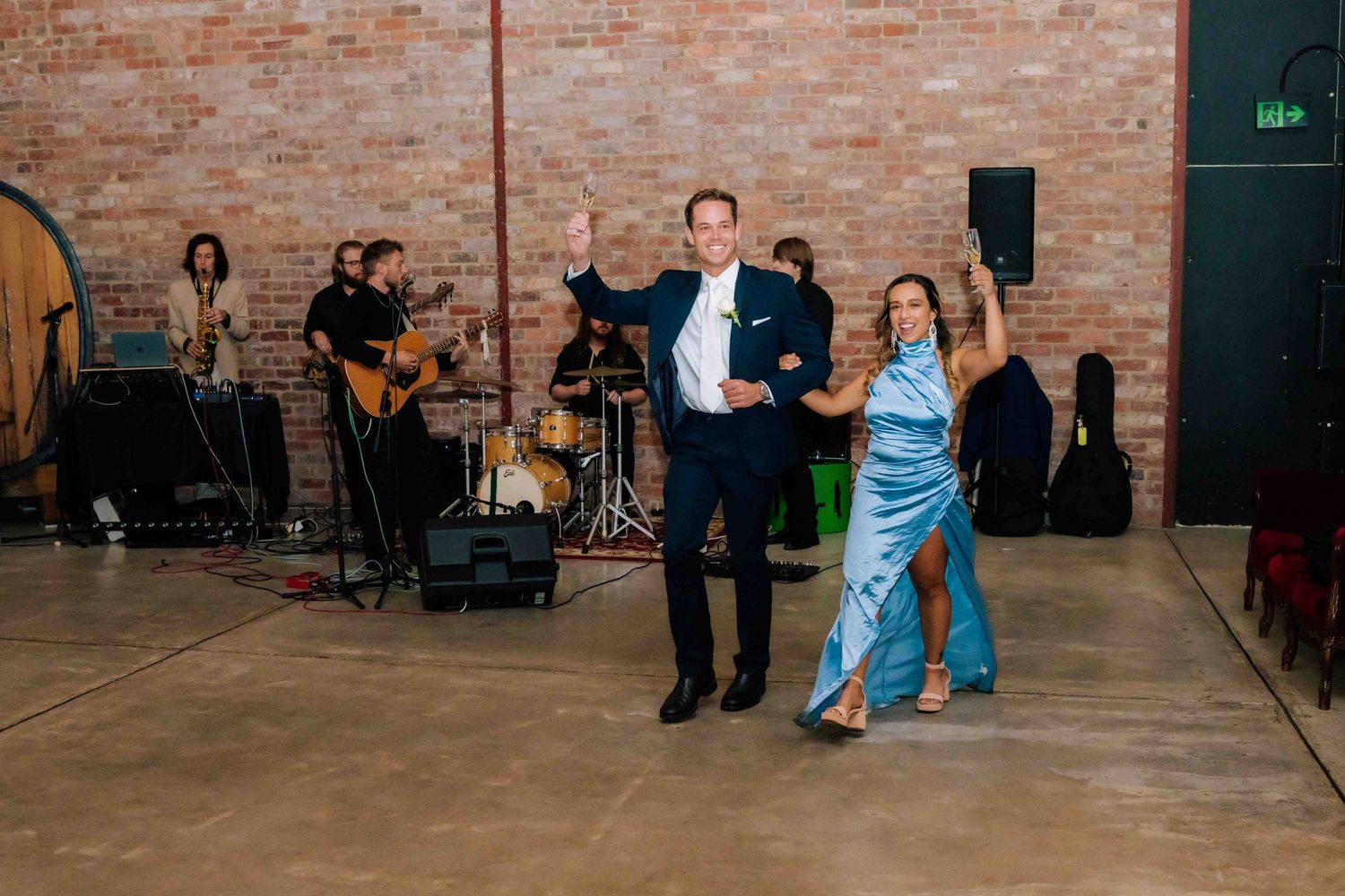 Couple dancing energetically on dance floor with live band performing in brick-walled wedding venue.