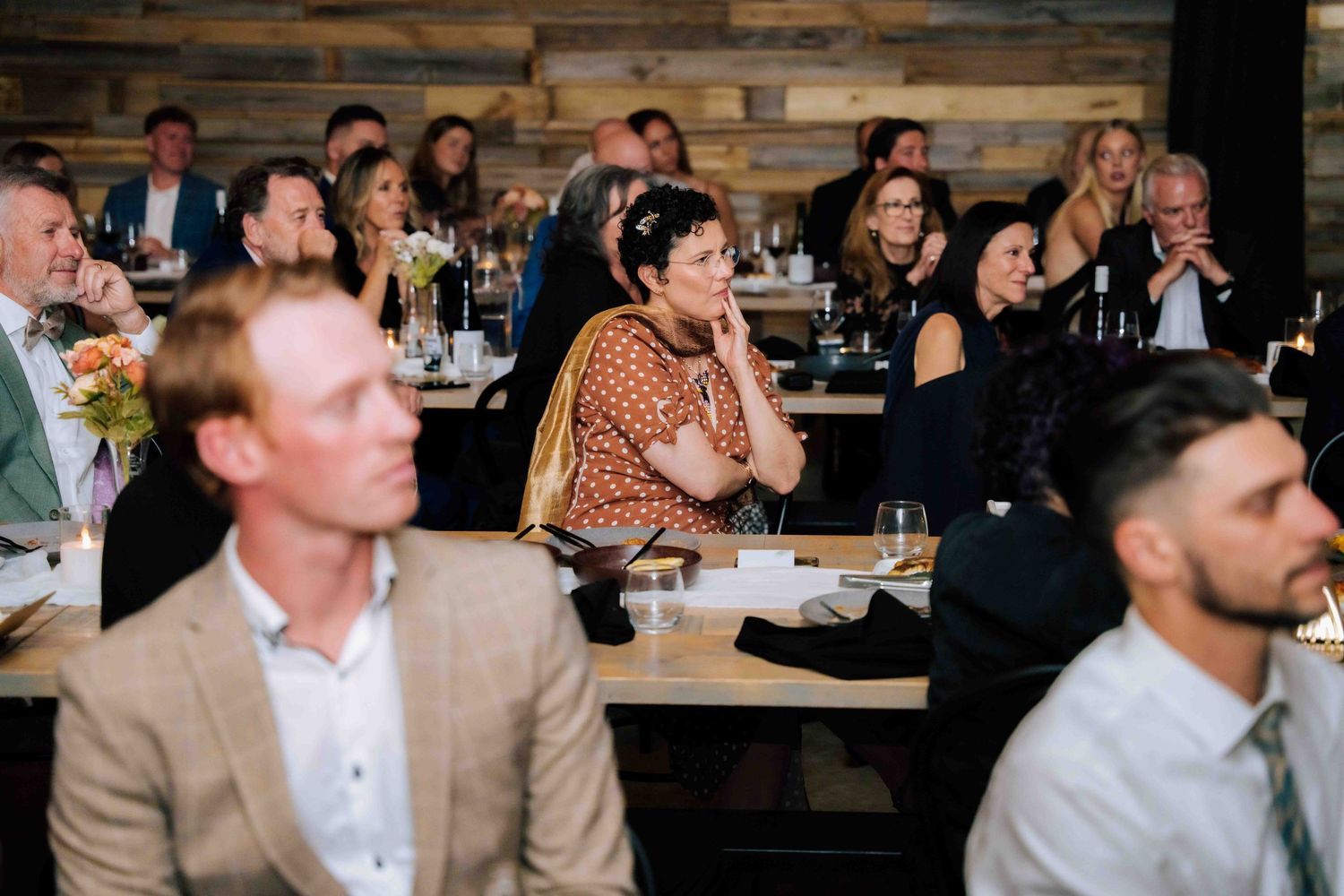 Wedding guests seated at tables attentively watching a speech during a formal reception dinner.